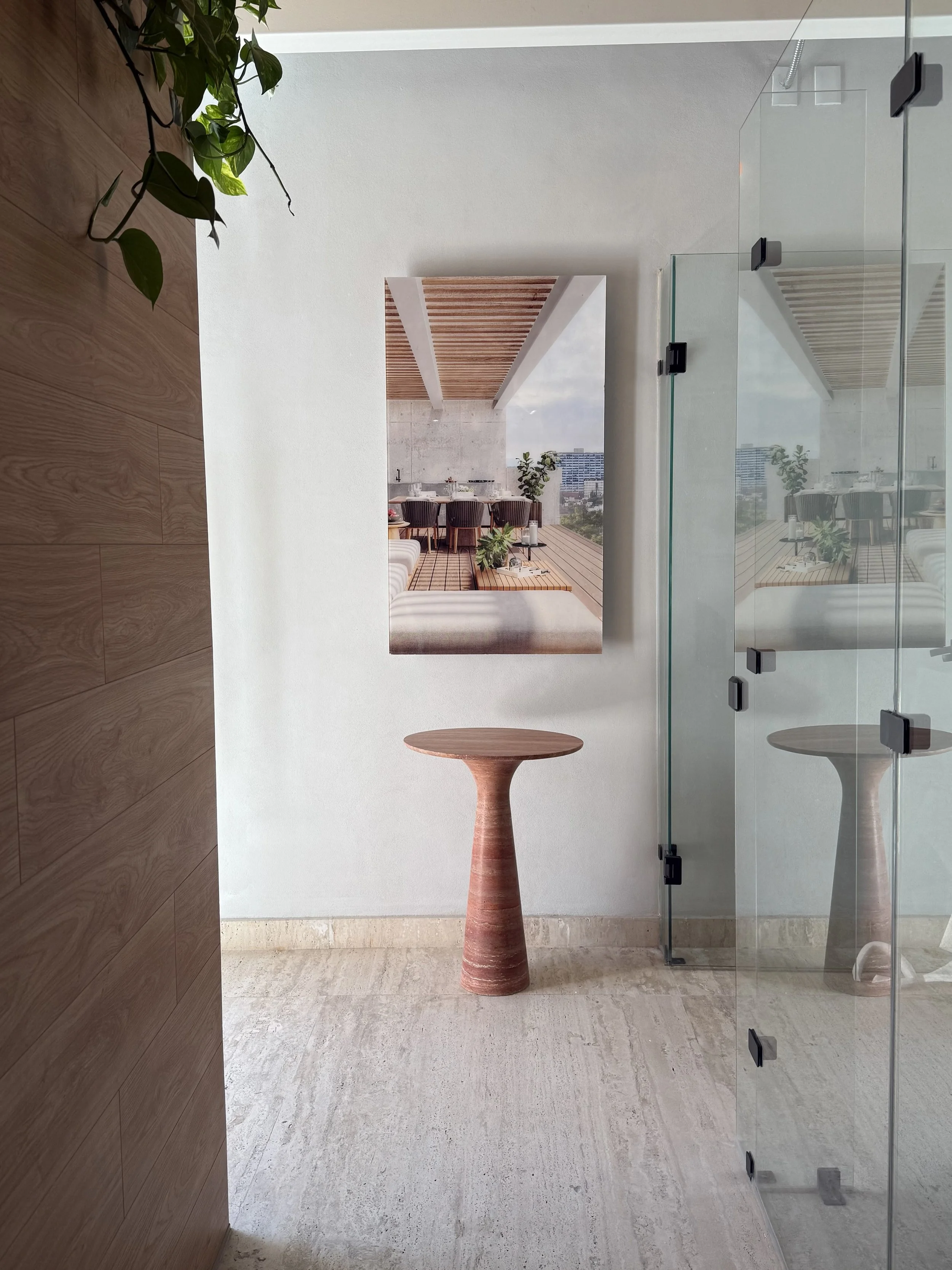 Vista de un interior minimalista con pared blanca, puerta de vidrio, un cuadro en la pared y una mesa de madera con base cilíndrica, reflejada en el vidrio.