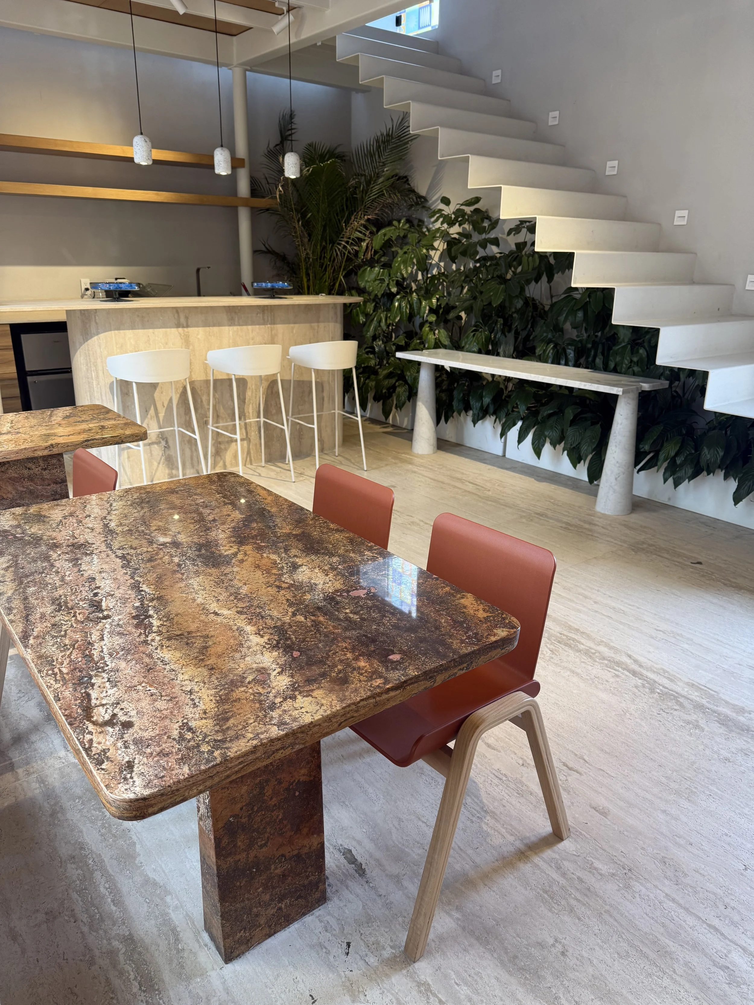 Interior moderno de una sala de estar con mesa de piedra, sillas de color terracota, barra con taburetes blancos, plantas verdes y una escalera blanca en el fondo.