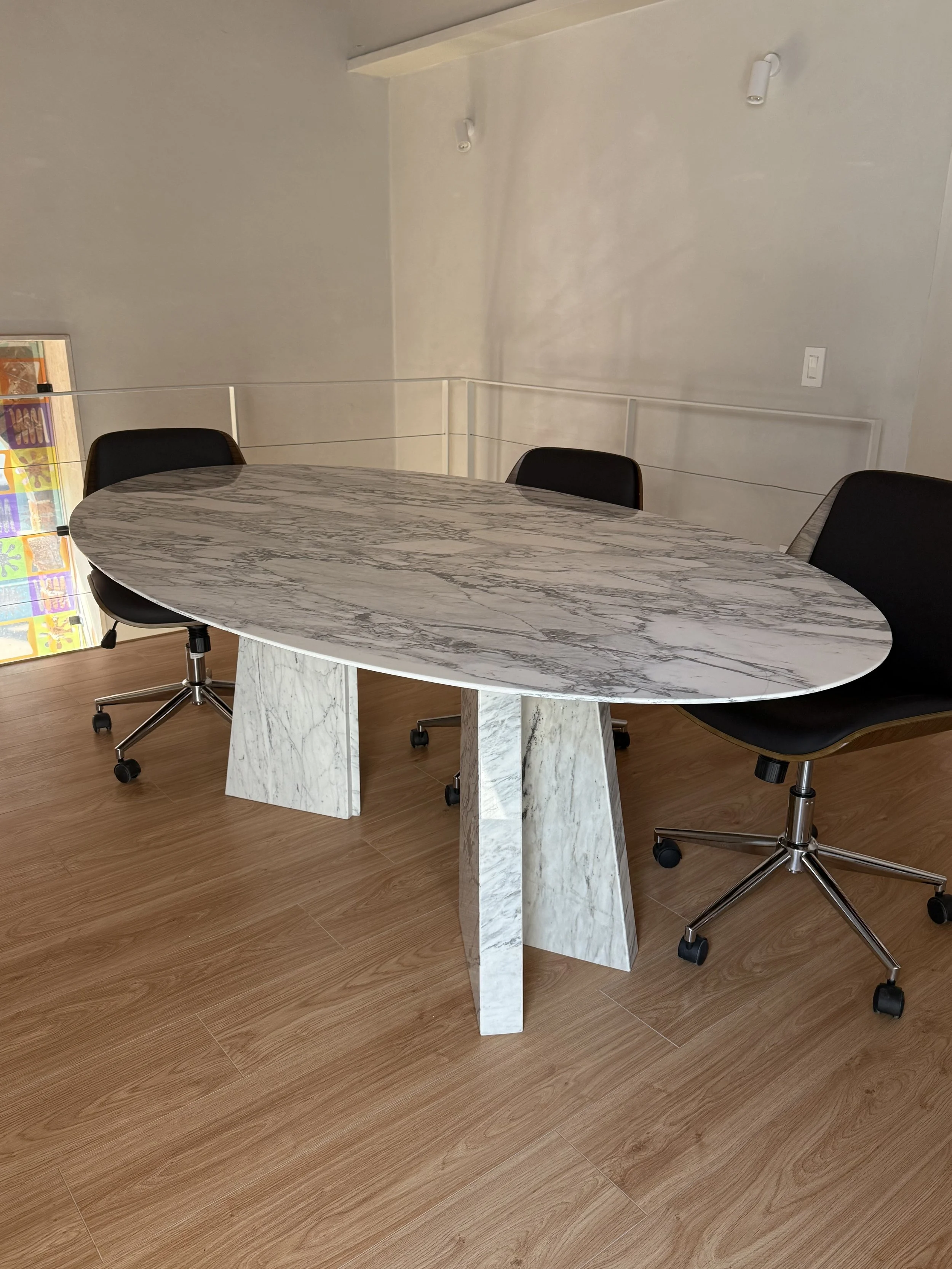 Mesa ovalada de mármol con tres patas y cuatro sillas negras con base metálica y ruedas en un espacio interior con paredes blancas y suelo de madera.