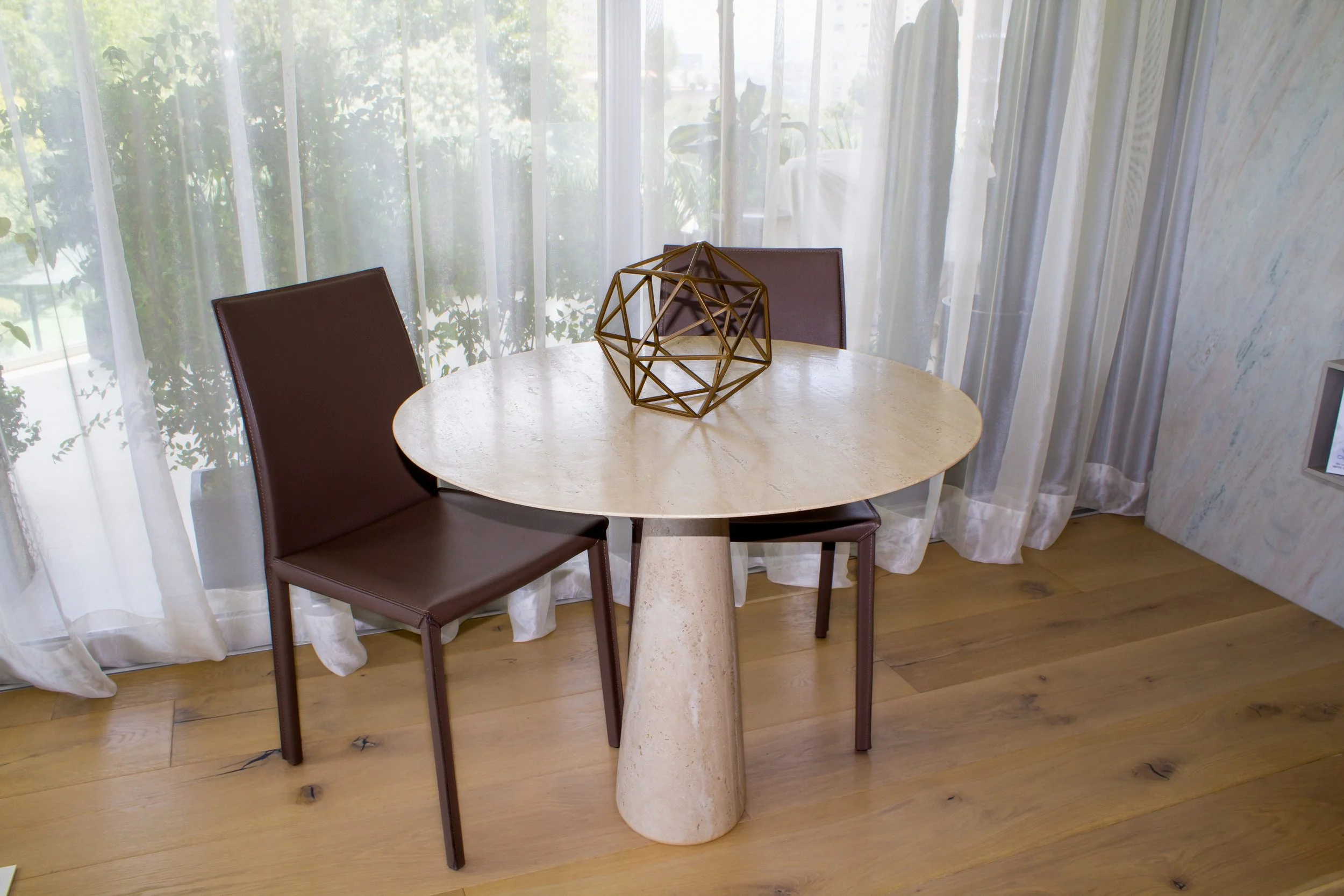 Mesa redonda de piedra natural travertino, para dos personas en una habitación con cortinas e iluminación natural y piso de madera.