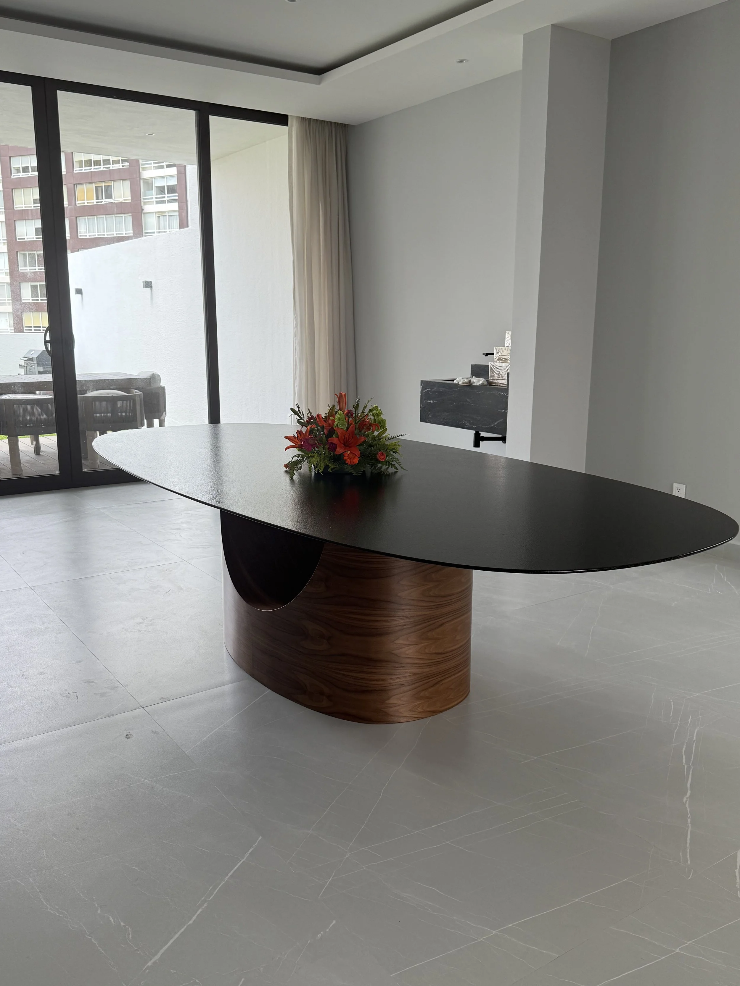 Mesa ovalada de color negro mármol monterrey con base de madera y un arreglo en el centro, en una habitación moderna con piso de cerámica, departamentos modernos, mobiliario de lujo.