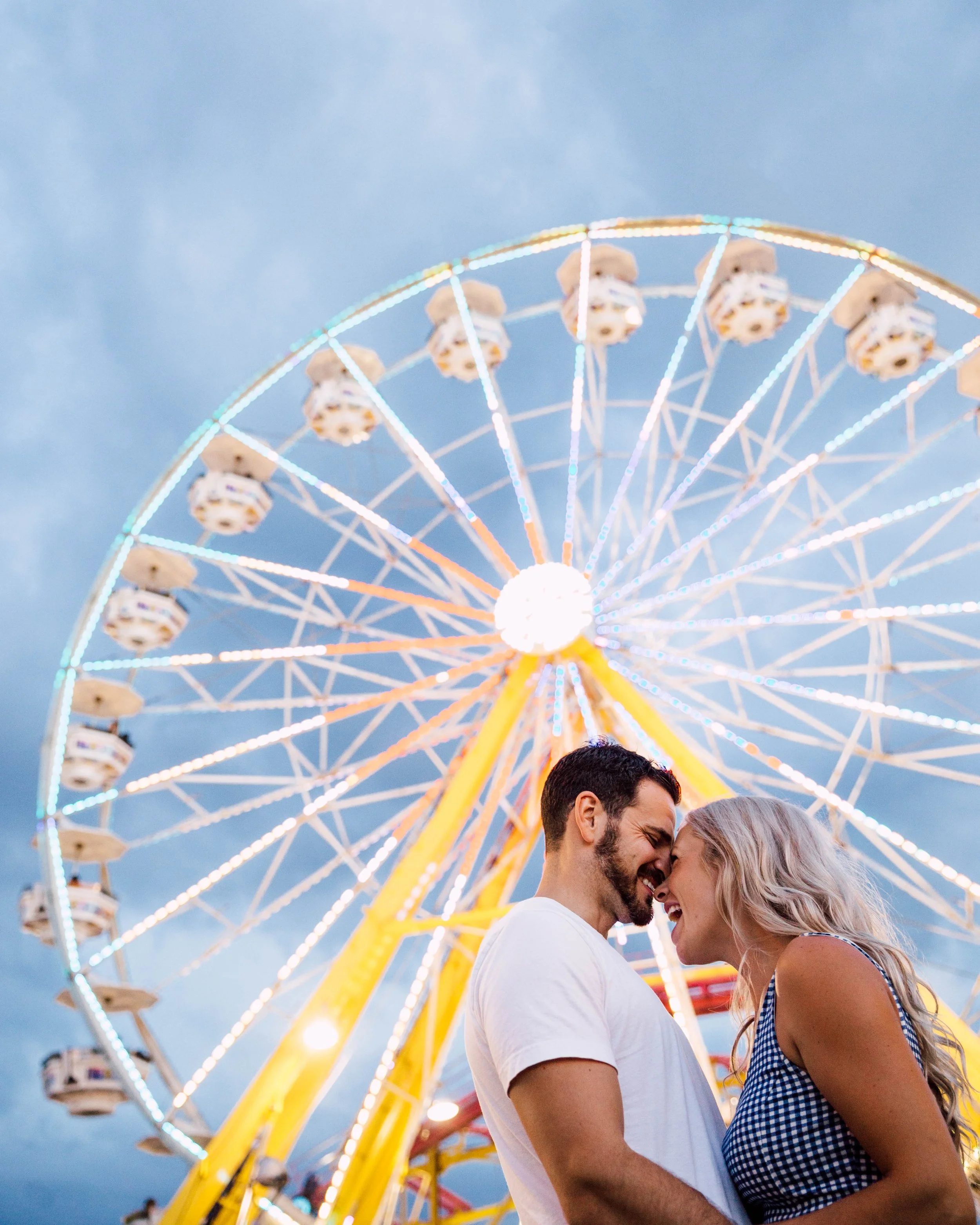 Couple smiling under a Ferris wheel at a fair.