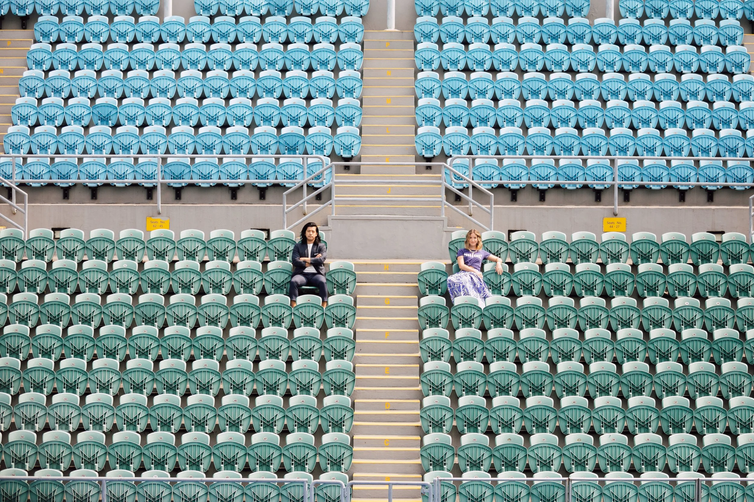 Two people sitting far apart in a mostly empty stadium with blue and green seats.