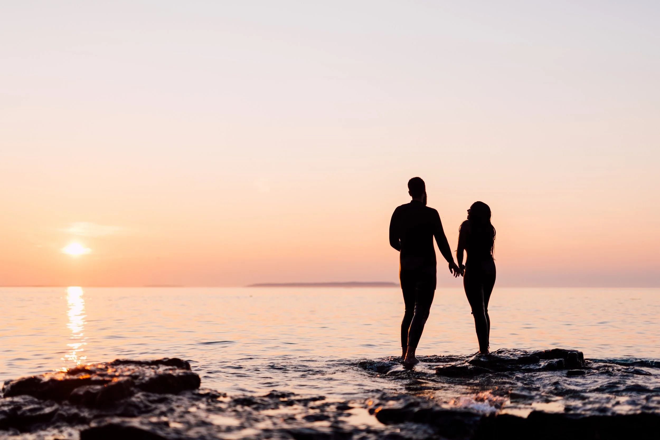 Silhouette of a couple holding hands standing on rocks by the sea during a sunset.