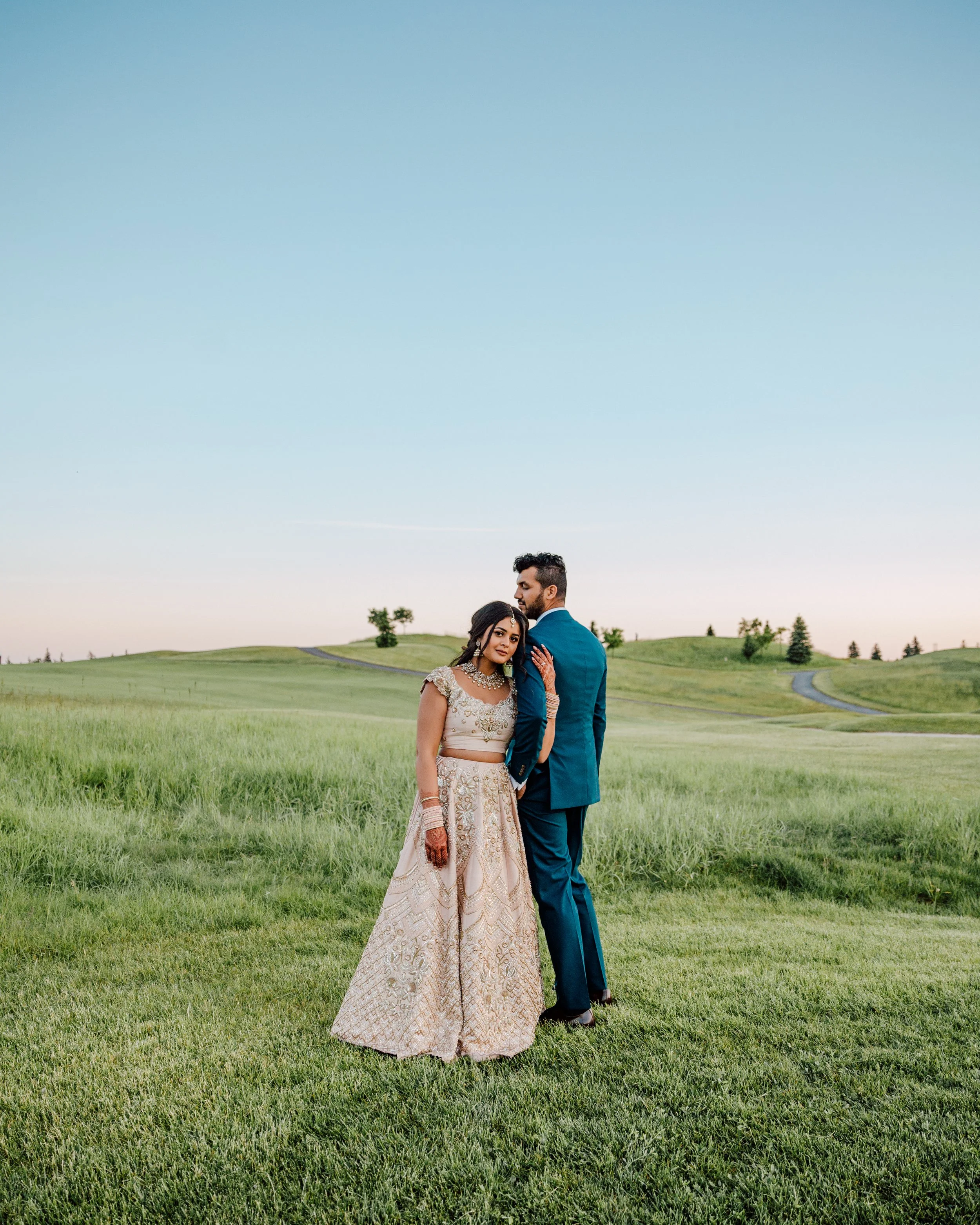A couple poses on a grassy landscape under a clear sky, with the woman wearing an embellished traditional dress and the man in a blue suit.
