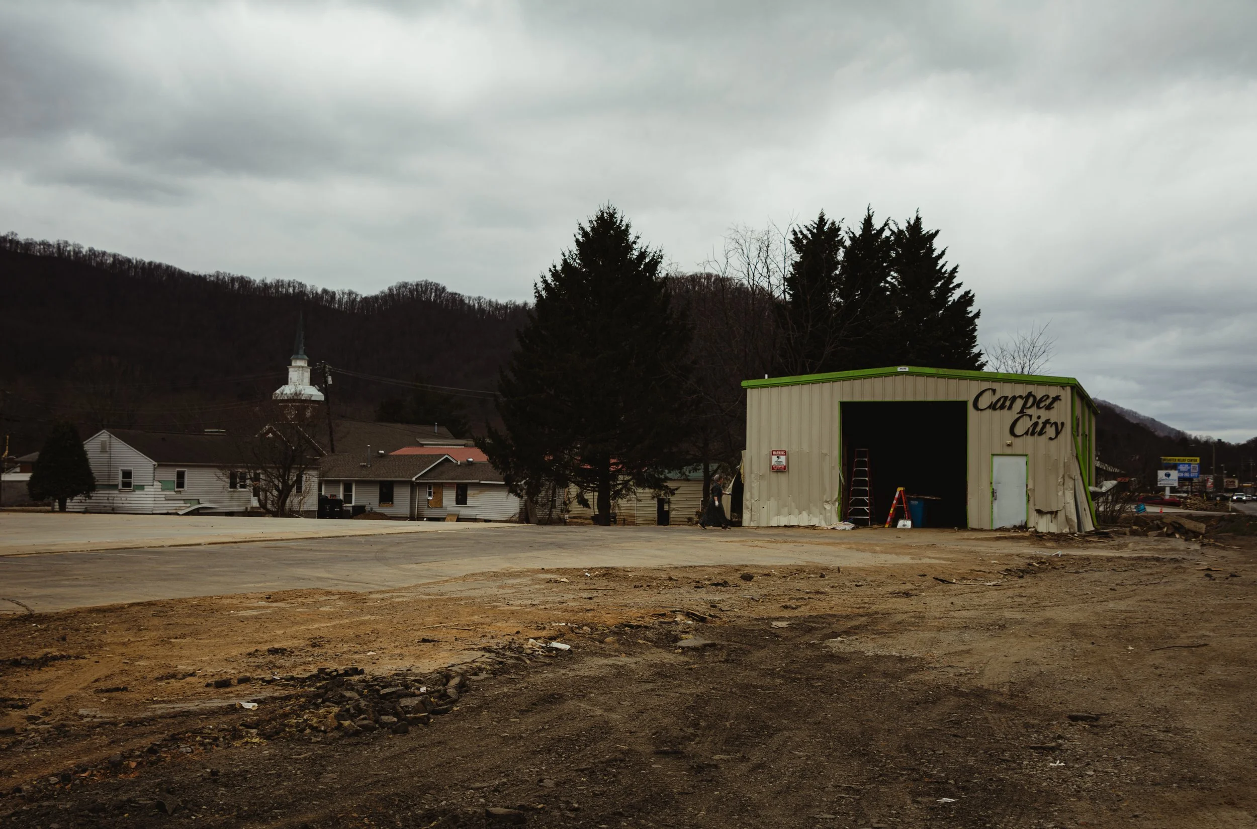 An overcast daytime scene with a small, damaged building labeled 'Carpet City' on the right, surrounded by dirt and debris. In the background, there are houses and a church steeple, with mountains and cloudy sky in the distance.