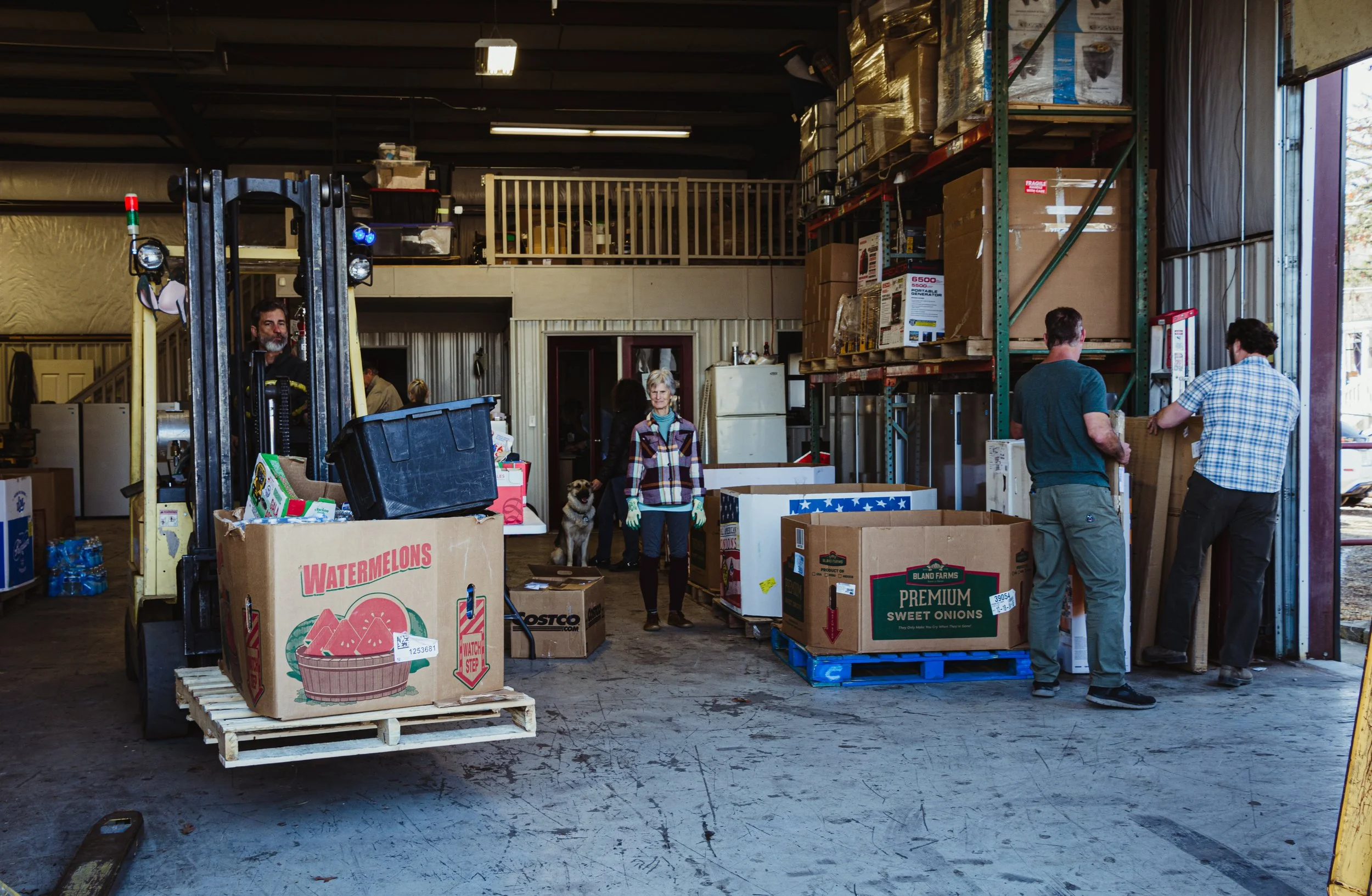 People working inside a warehouse or storage unit, handling boxes and supplies, with shelves stocked with items and a forklift.