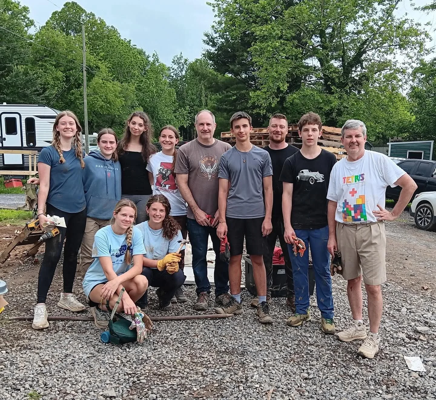 Group of eleven people, including young teens and adults, standing outdoors on a gravel surface with trees and parked cars in the background. Some are holding gloves and tools, indicating a volunteer or community service activity.