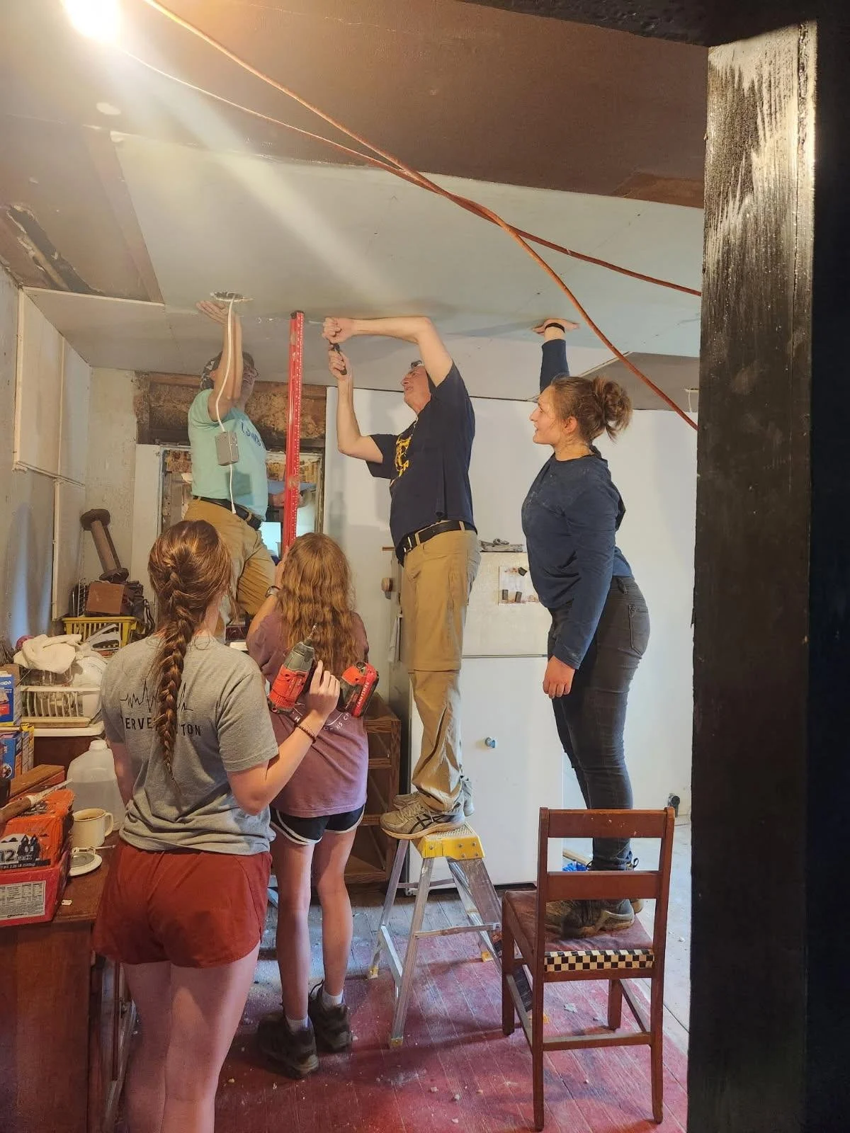 A group of five people working together to install drywall on a ceiling in a room, with some standing on chairs and a ladder, using tools and measuring tape.