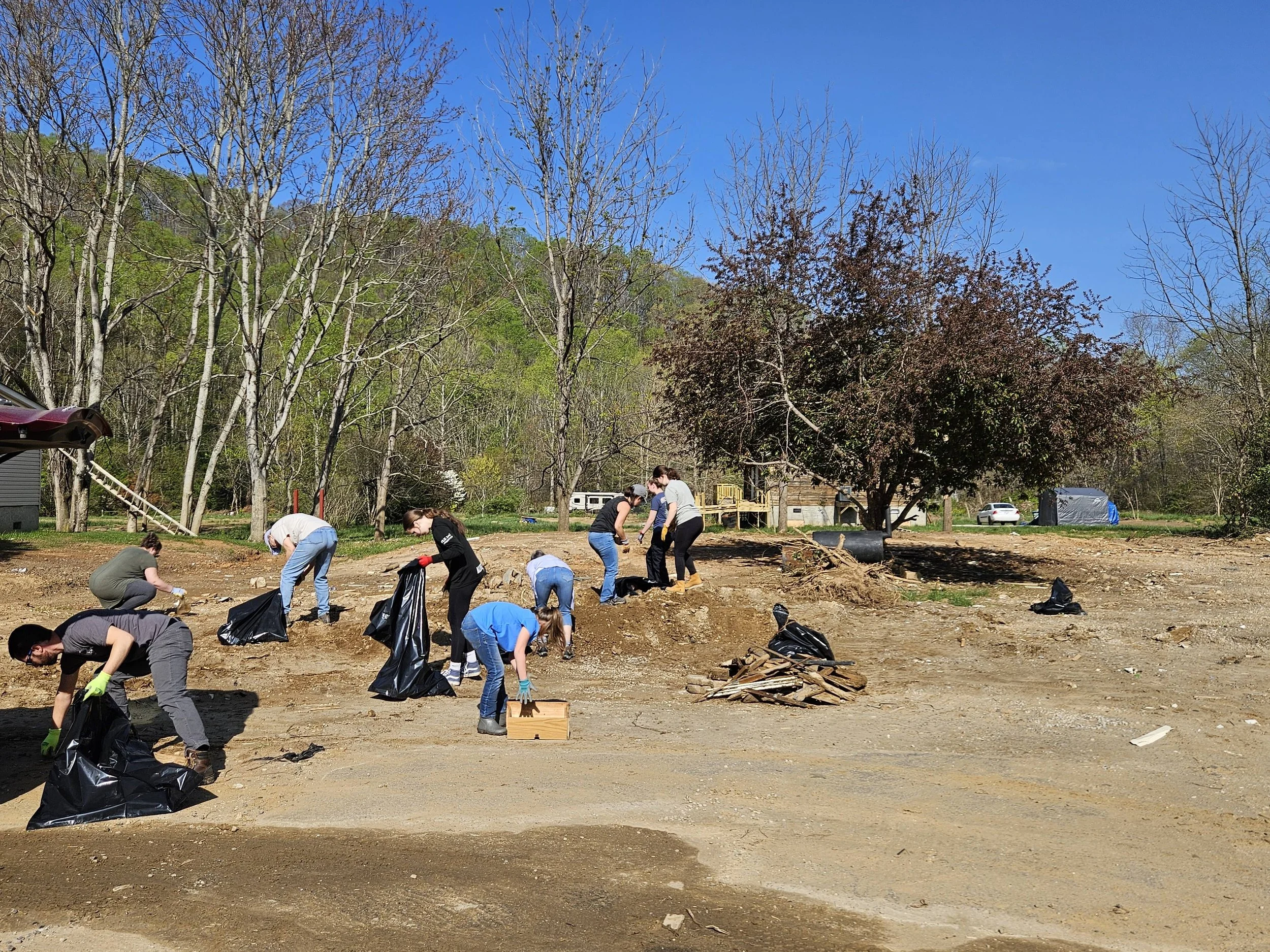 People participating in a community clean-up, picking up trash and placing it in large black garbage bags in an outdoor setting with trees and a clear blue sky.
