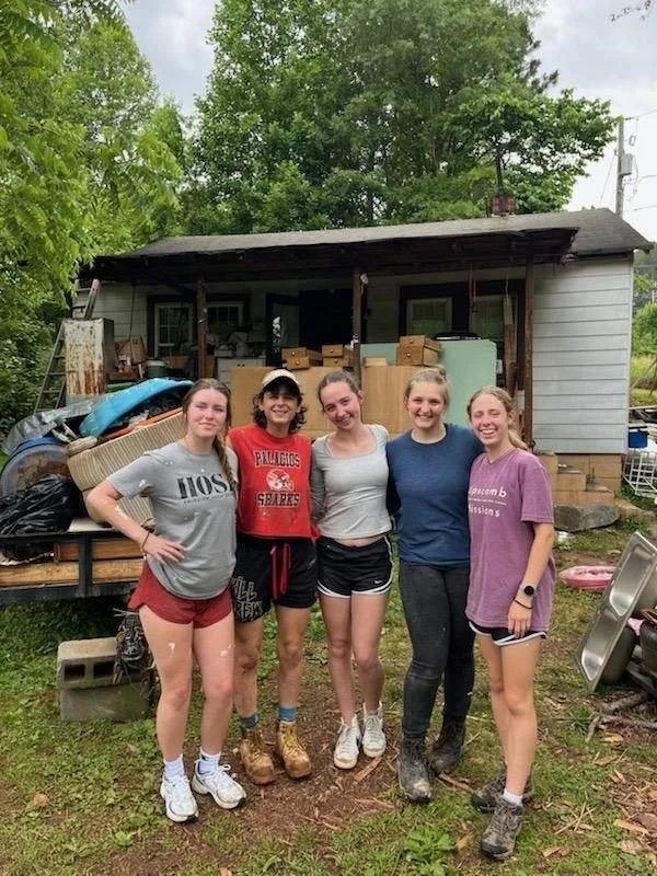 Five young women stand together in front of a cluttered house or shed with various items stacked on the porch, surrounded by green trees.