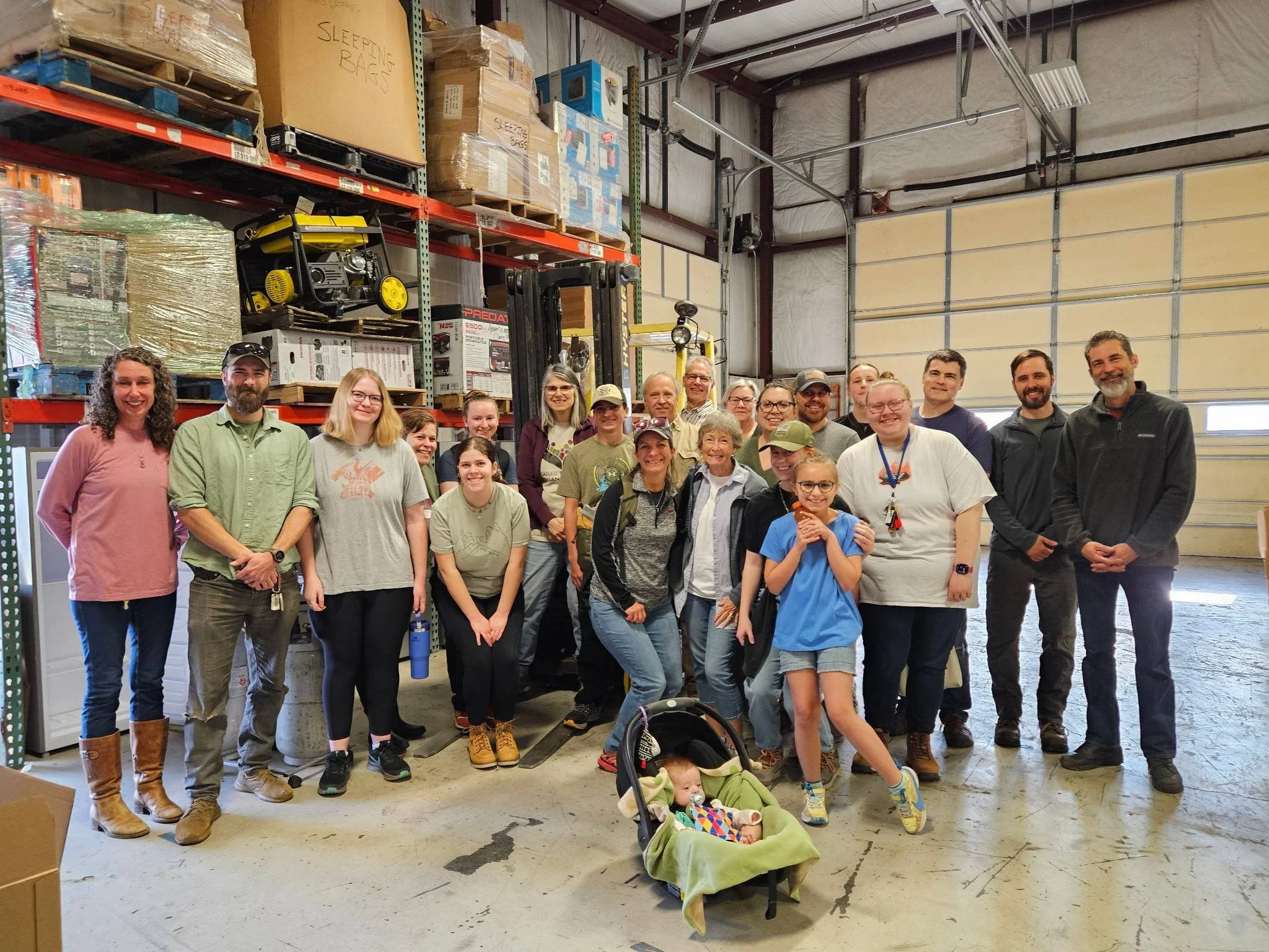 A large group of people, including adults and children, posing for a photo inside a warehouse with shelves of boxes and equipment in the background, some smiling and others making playful gestures.
