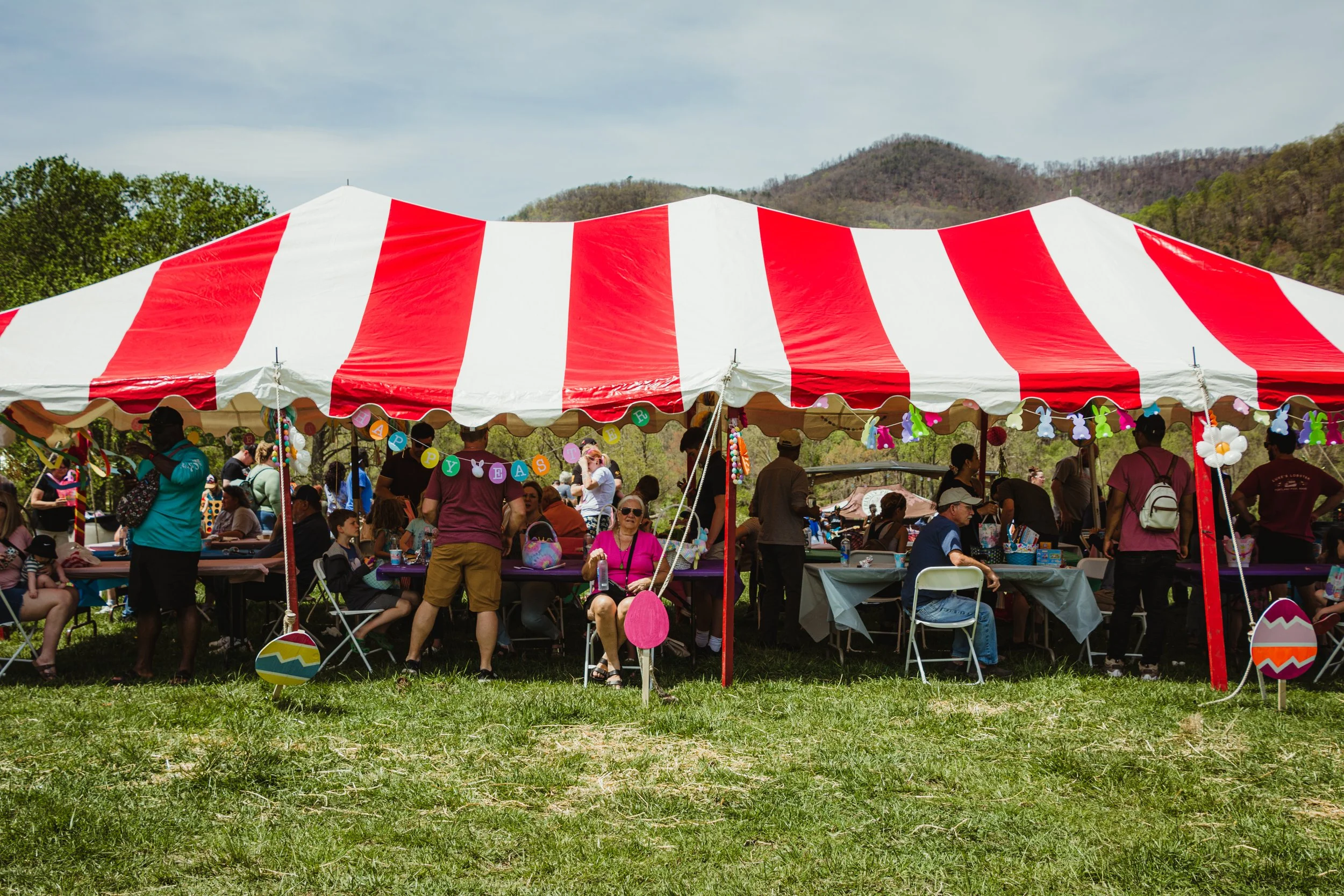 Outdoor Easter celebration with a large red and white striped tent, decorated with Easter-themed banners and hanging decorations, set against a backdrop of mountains and greenery.