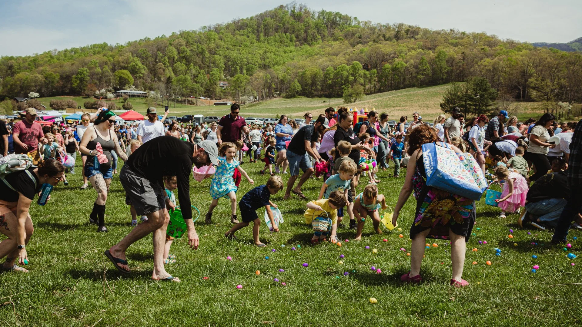 Large group of people, including children, participating in an Easter egg hunt on a grassy field with hills and trees in the background.