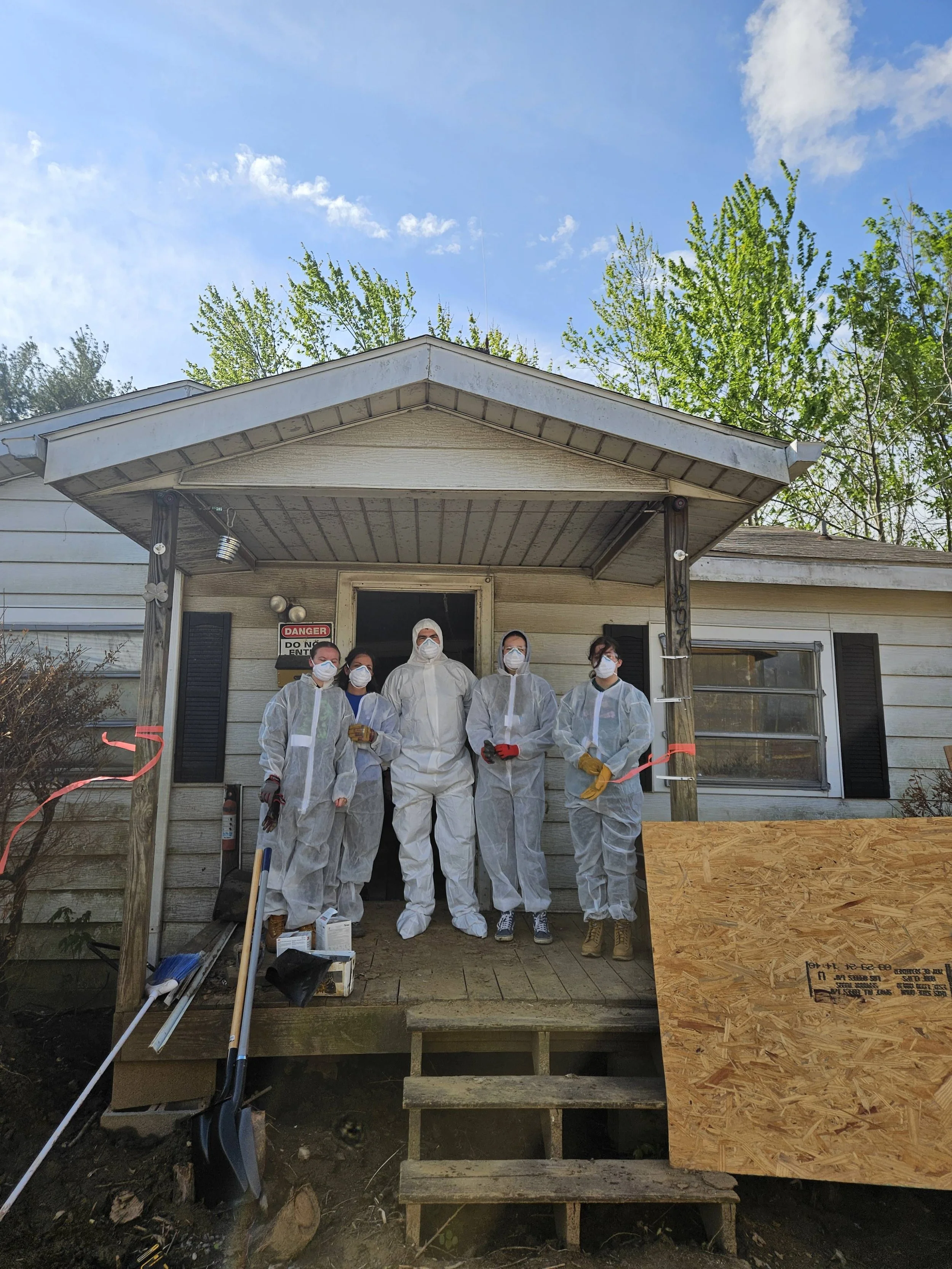 Six people wearing protective gear and masks standing in front of a house with construction materials around, possibly during a construction or renovation project.