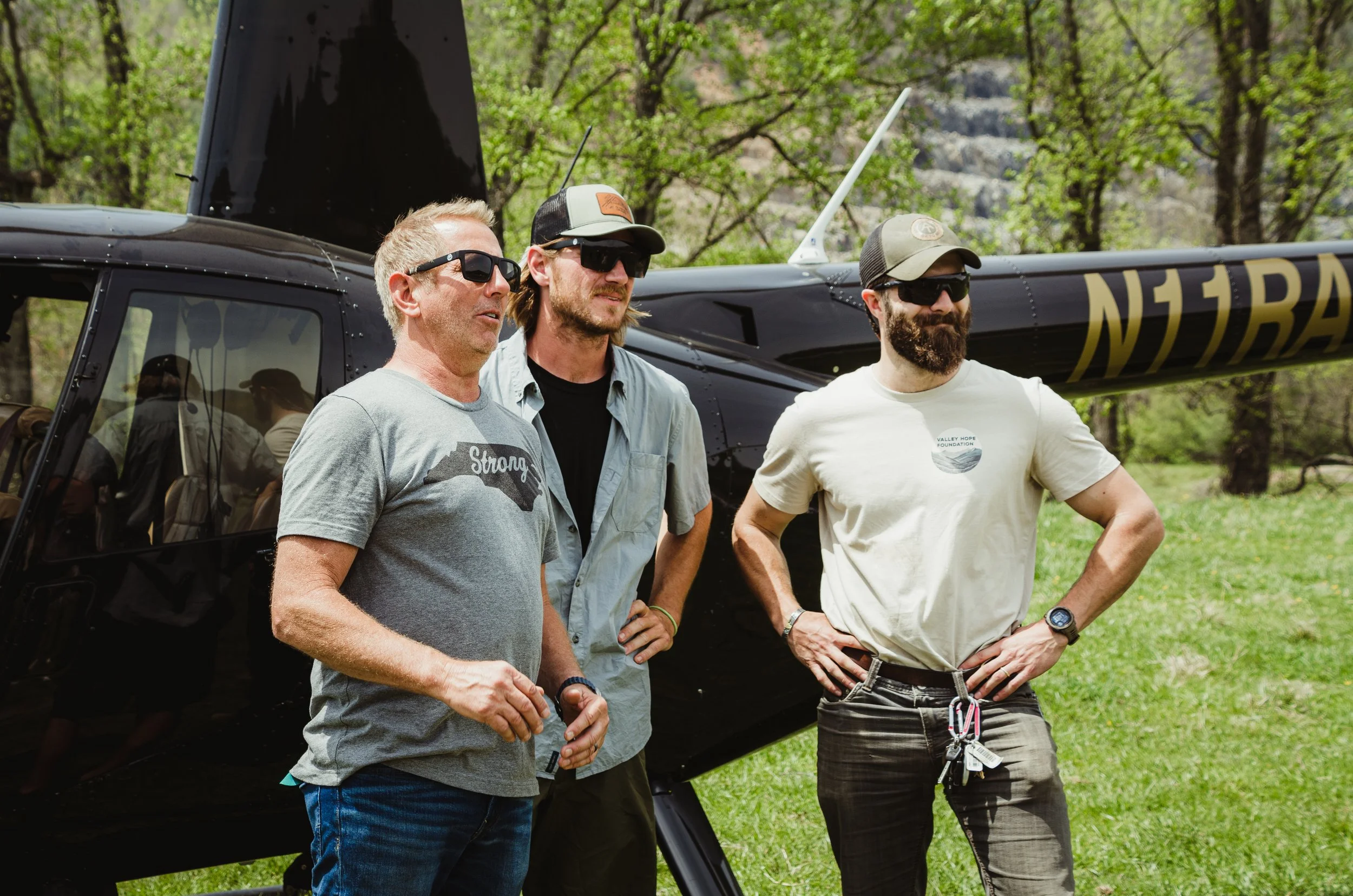 Three men standing outdoors in front of a helicopter with trees and greenery in the background. They are wearing sunglasses and casual clothes.