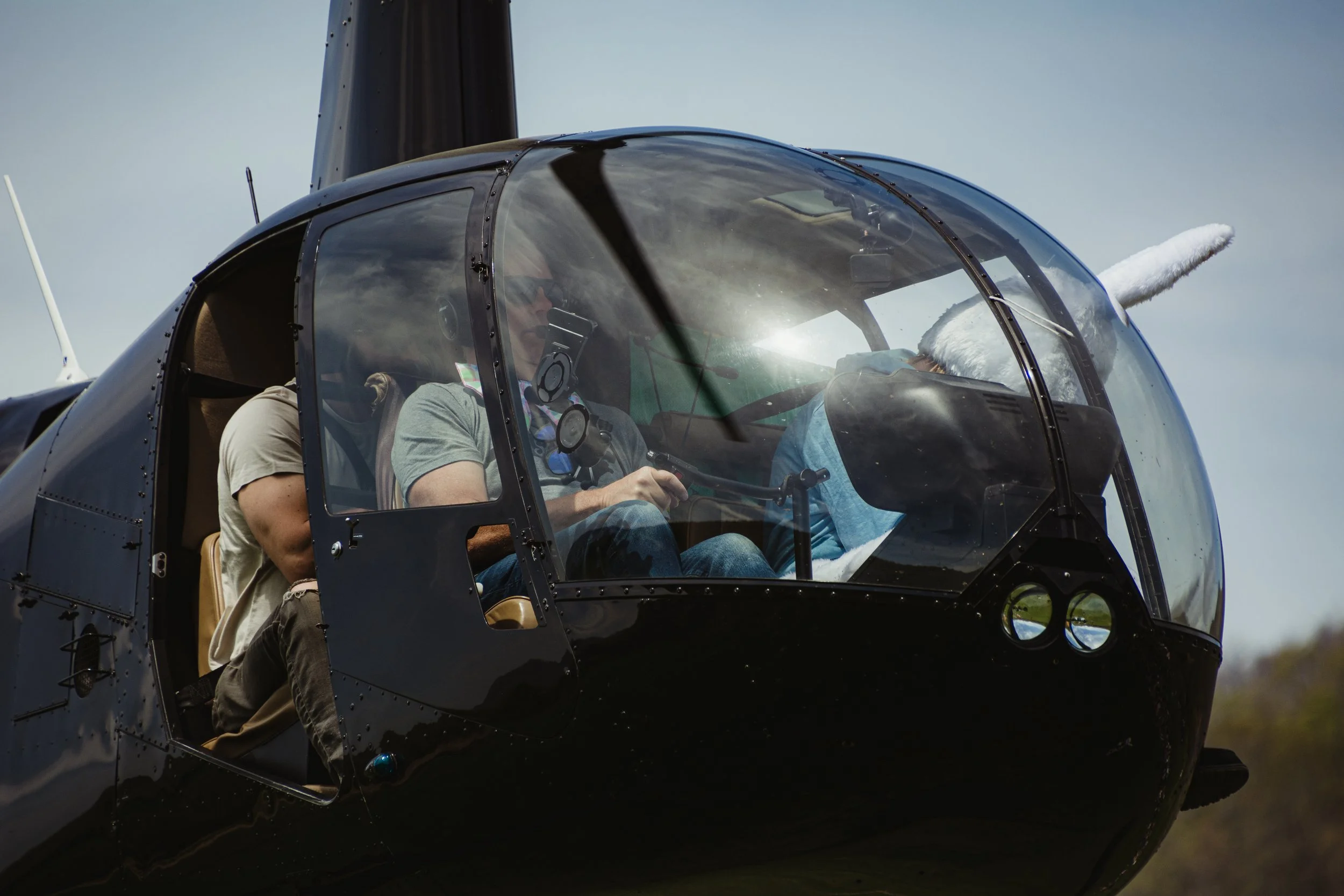 Close-up of the cockpit of a helicopter with two people inside, one person is wearing sunglasses and a headset, appearing to operate the controls.