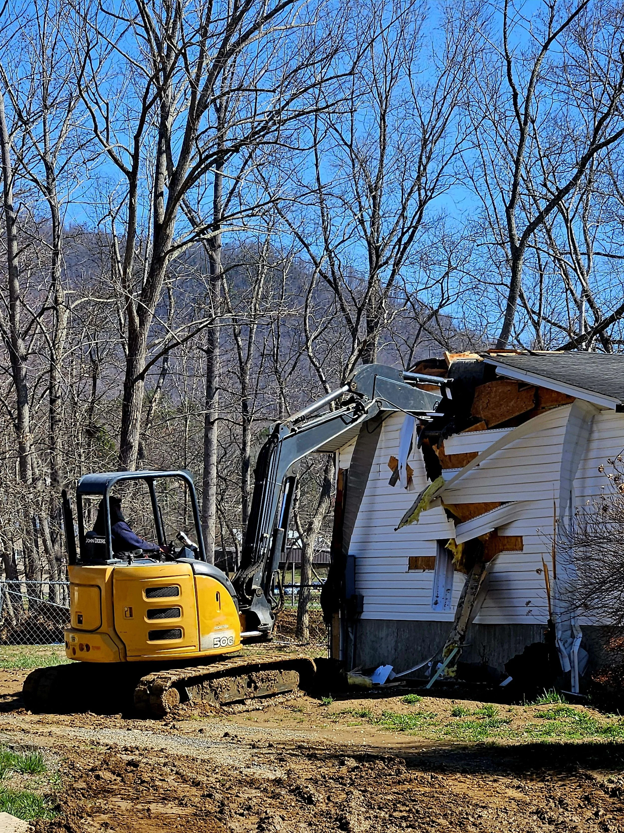 A small yellow excavator demolishing a white house with a partially collapsed roof on a clear day.