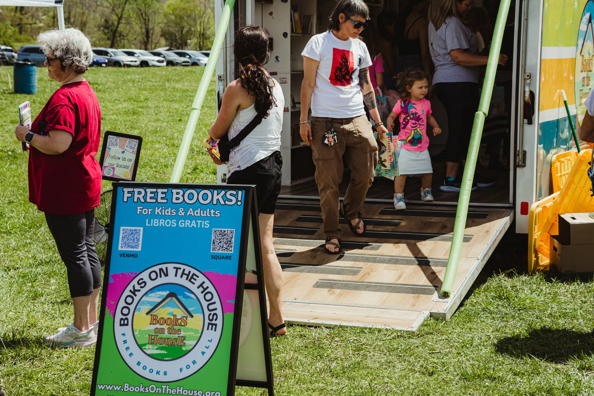 A group of people, including children, entering a mobile book fair titled 'Books on the House' with a sign advertising free books for kids and adults, set up on a grassy outdoor area on a sunny day.