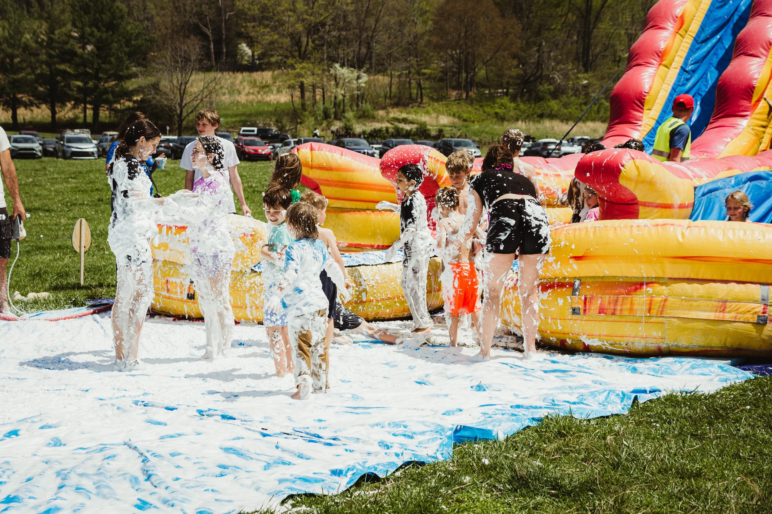 Children playing on a foam slip and slide with foam and water, outdoors on grass, with a yellow and red inflatable slide and parked cars in the background.