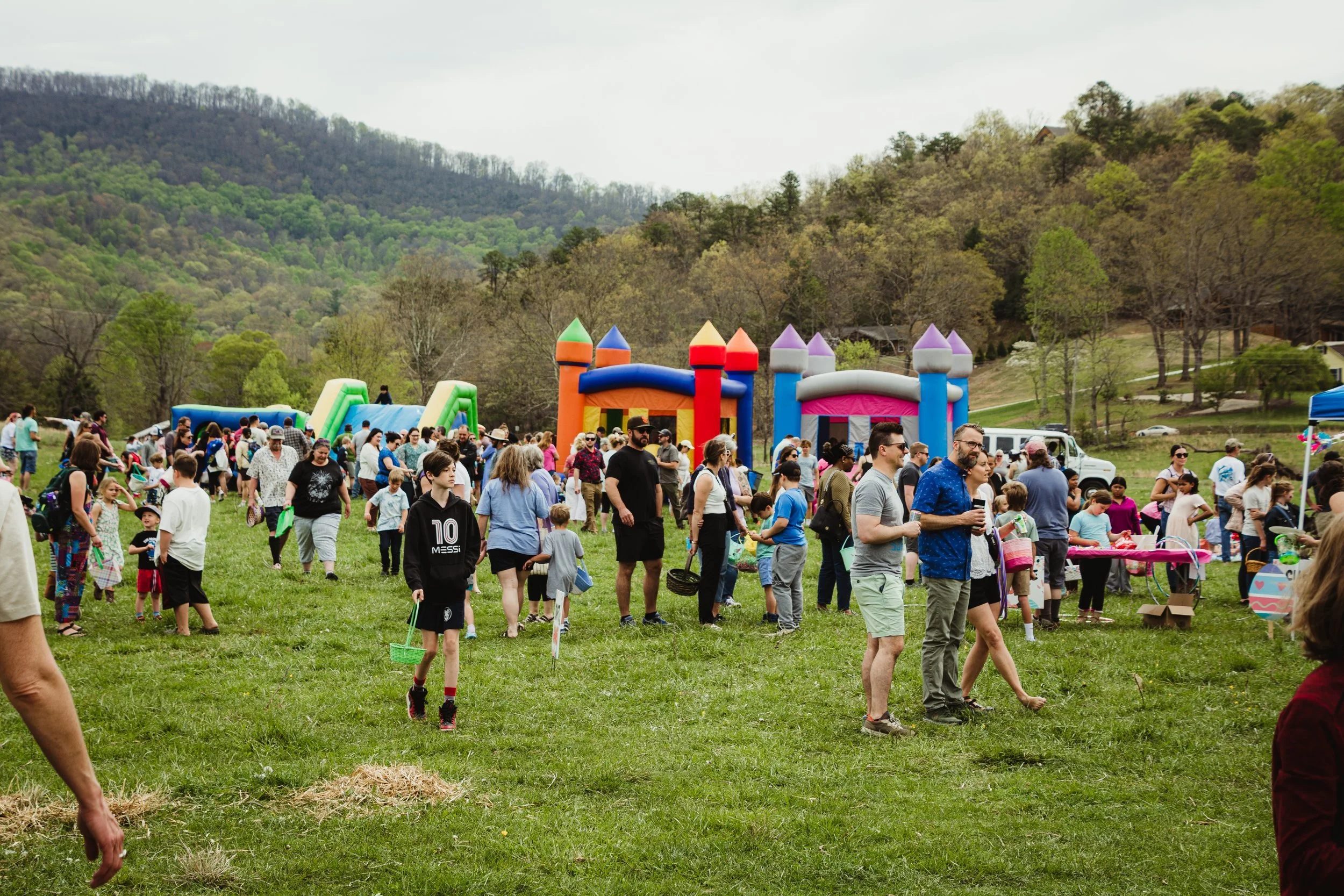 People at an outdoor event with inflatable castles, surrounded by trees and mountains.