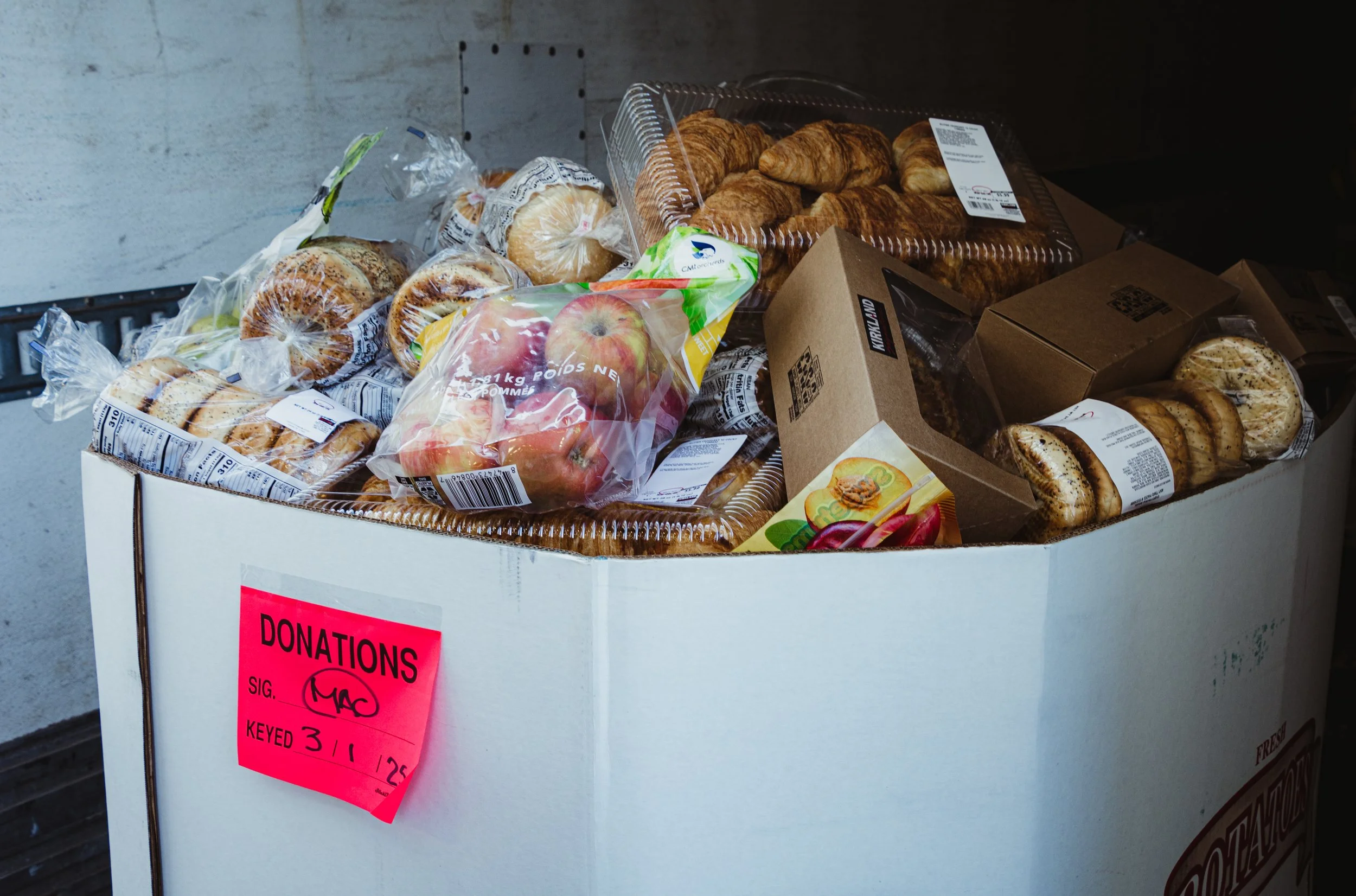A donation box filled with various baked goods and a bag of apples, with a pink donation label attached to the side.
