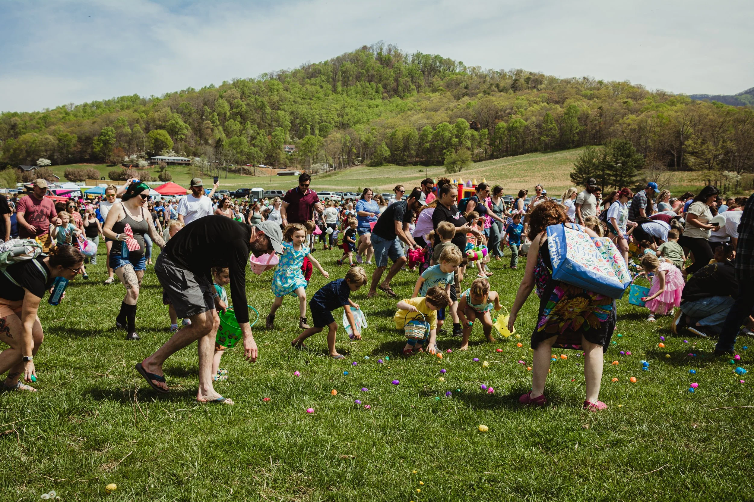 People, including children, participating in an Easter egg hunt on a grassy field with a mountain and trees in the background.