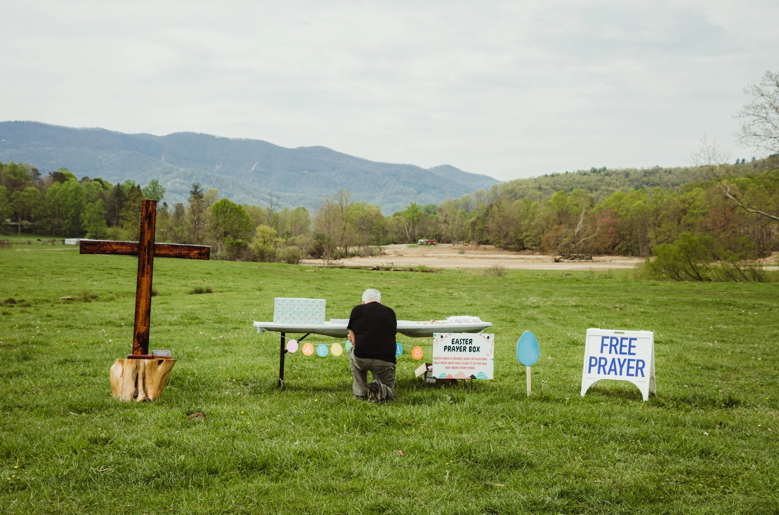 A person kneeling in prayer at an Easter outdoor prayer station with a large wooden cross on the left, a table with an Easter banner, a sign offering free prayer, and a large blue egg decoration set up on a grassy field with mountains and trees in th