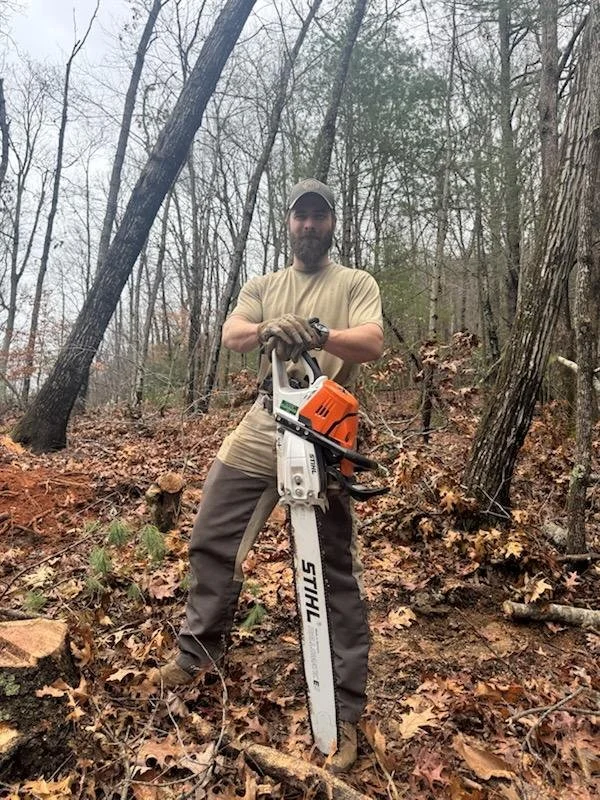 A man standing in a wooded area holding a chainsaw.