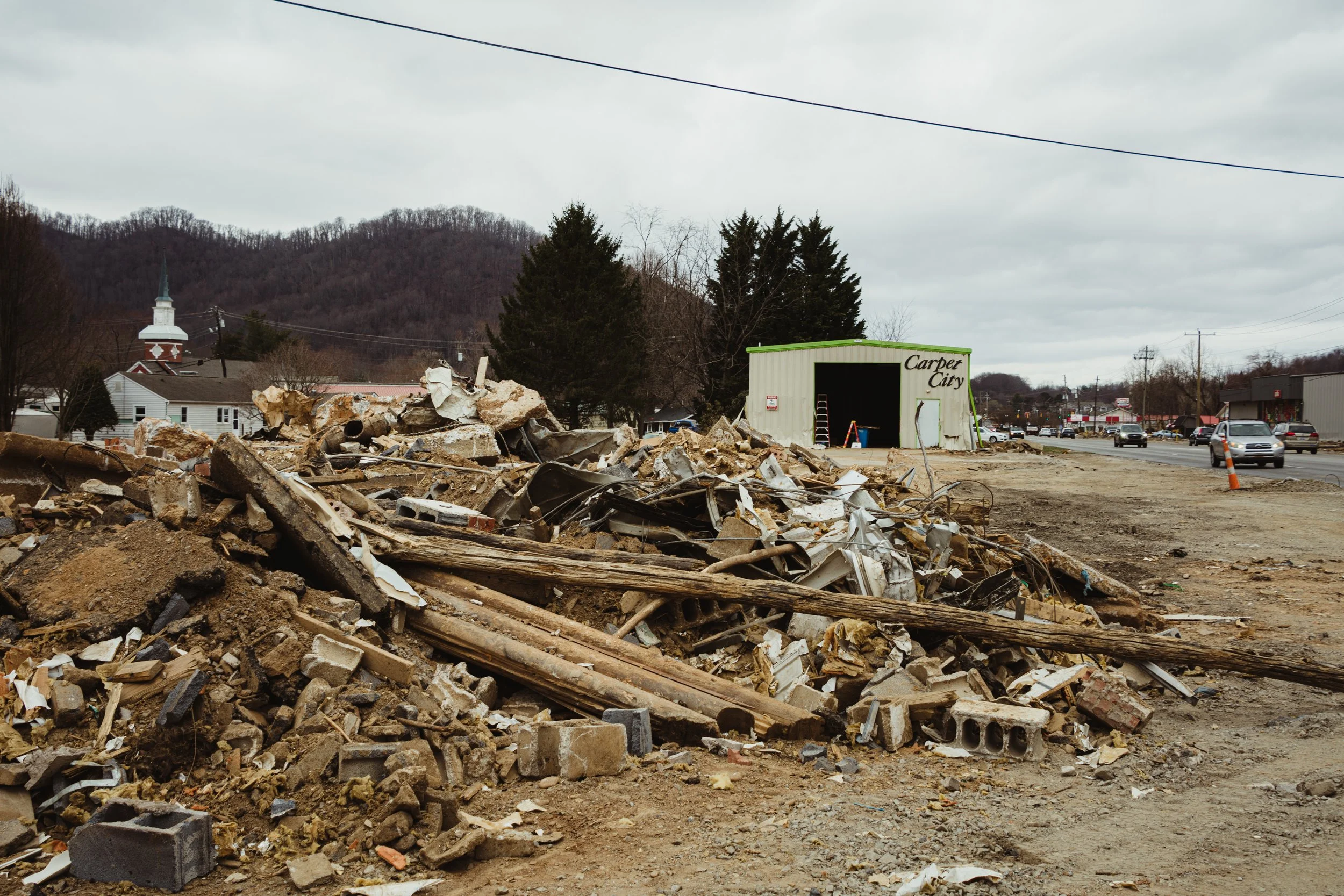 Debris and rubble from a construction or demolition site with a building labeled 'Carpet City' in the background, parked cars, and mountains in the distance under a cloudy sky.
