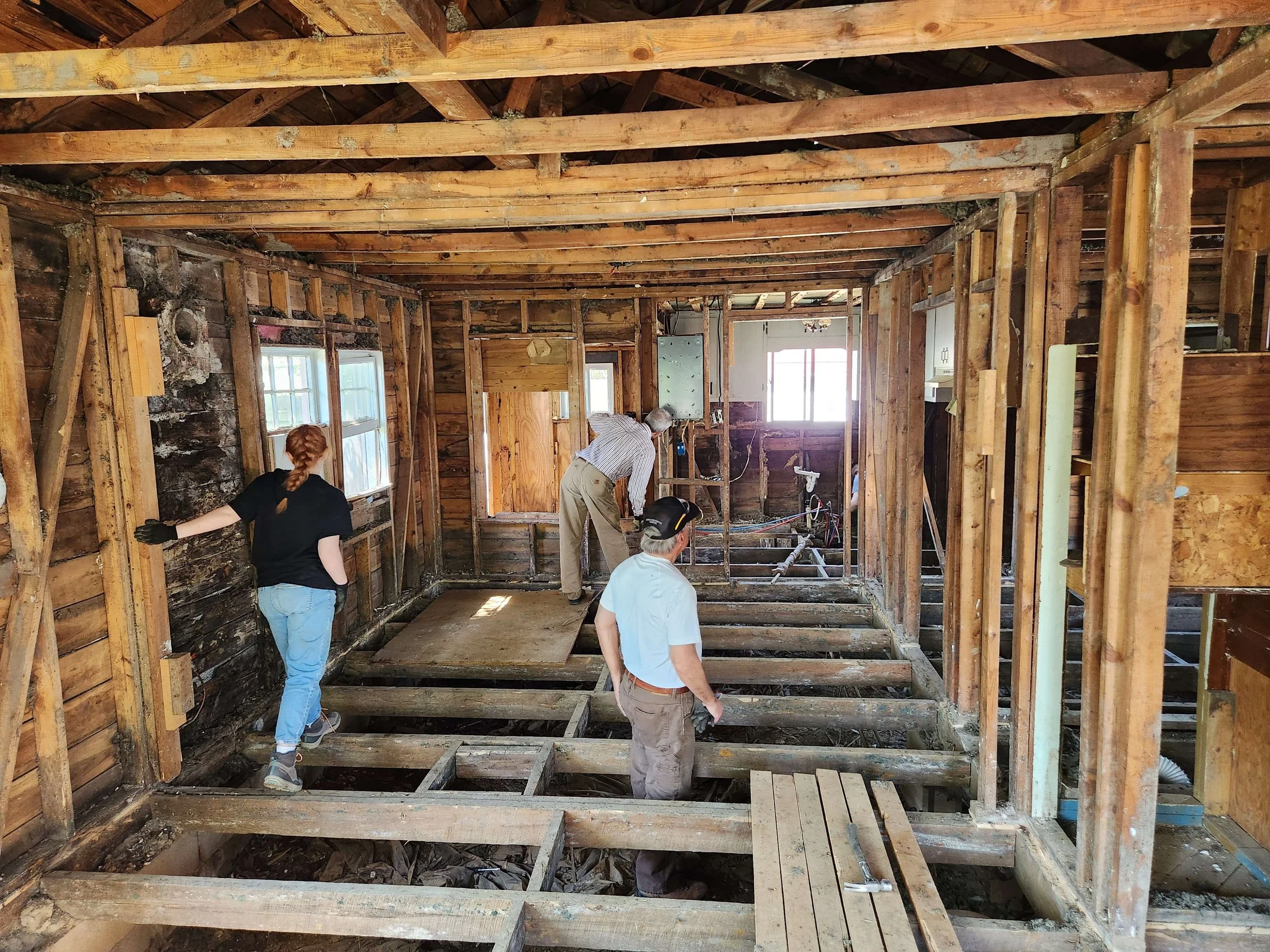People working on a house renovation, exposing wooden floor framing and walls, with some windows and an open door at the back.