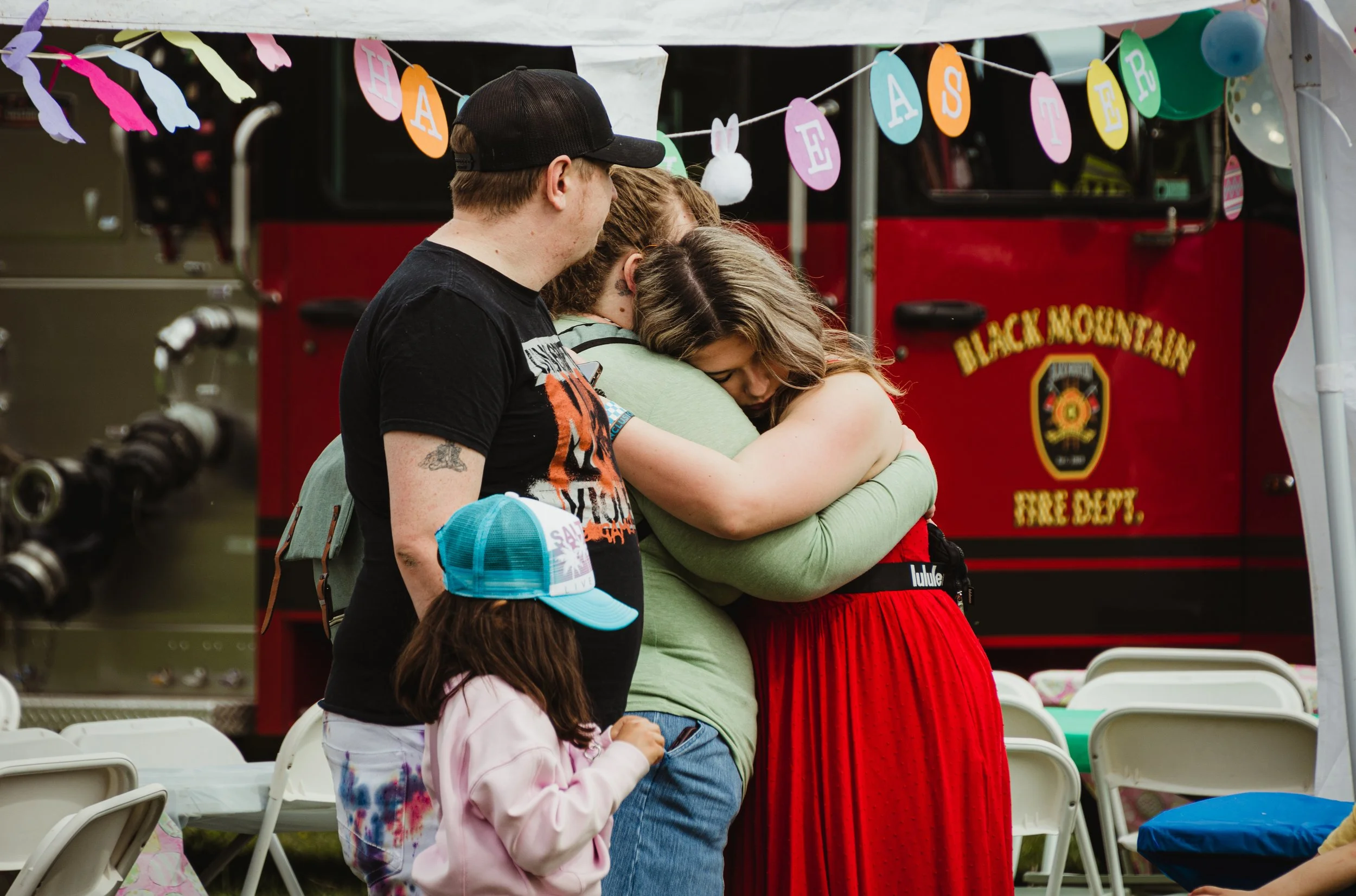 A group of people hugging at an outdoor event, with a fire truck in the background and pink, green, and yellow banners hanging above them.