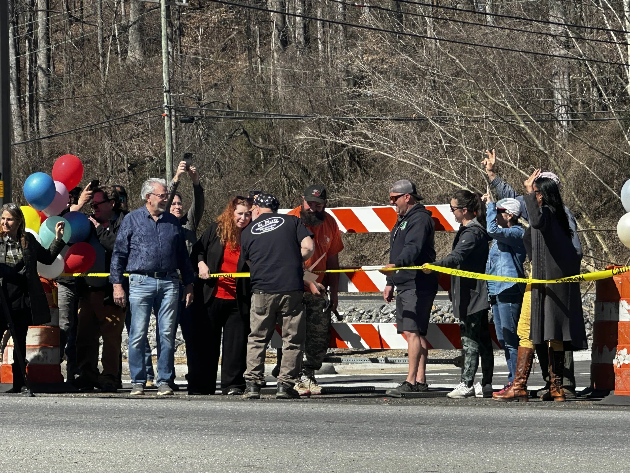 Group of people gathered at a road construction site, some holding balloons, taking photos, and celebrating, with yellow caution tape, construction barricades, and a wooded background.