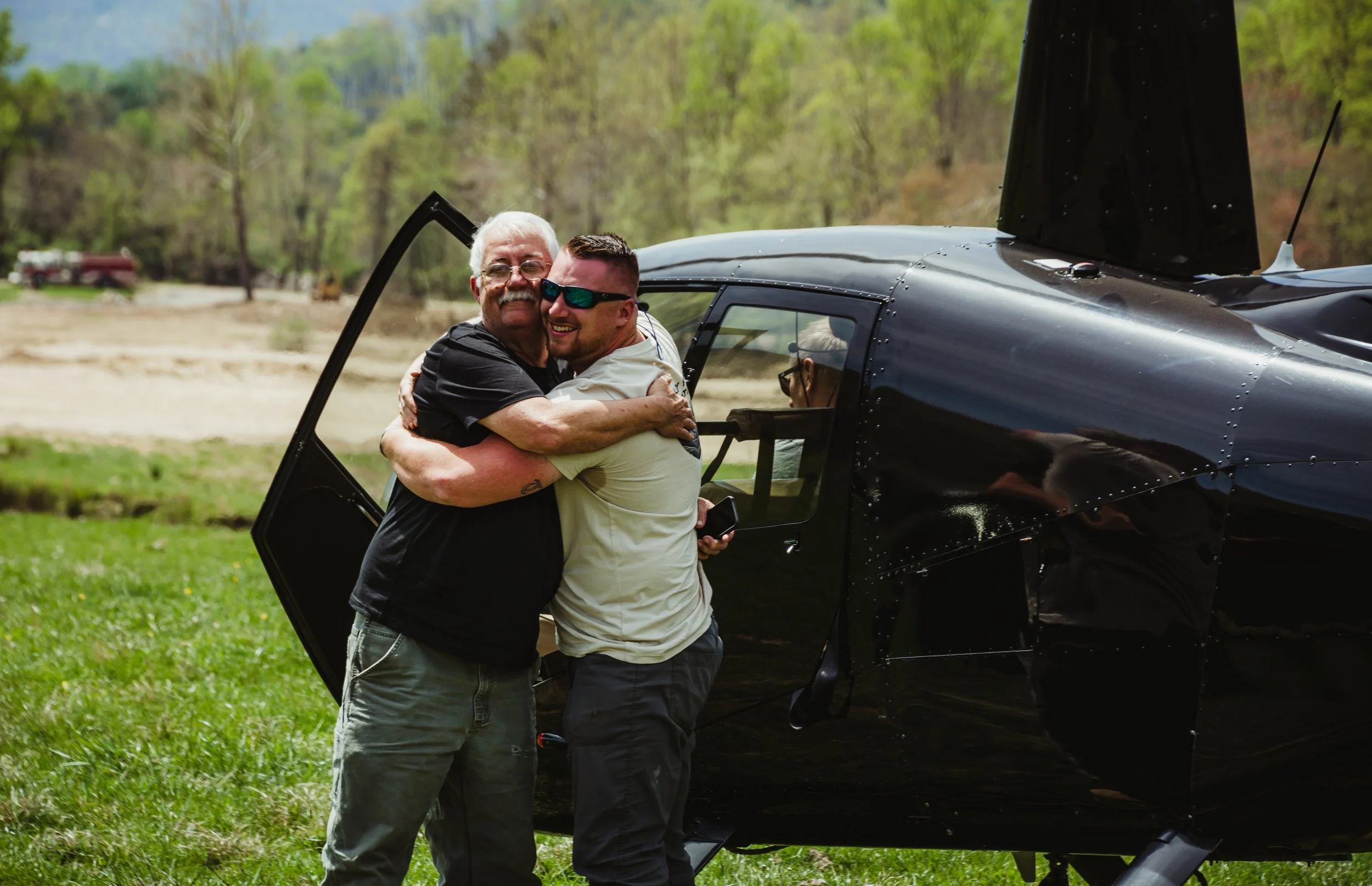 Two men hugging in front of a black helicopter on a grassy area, with trees and a partly cloudy sky in the background.