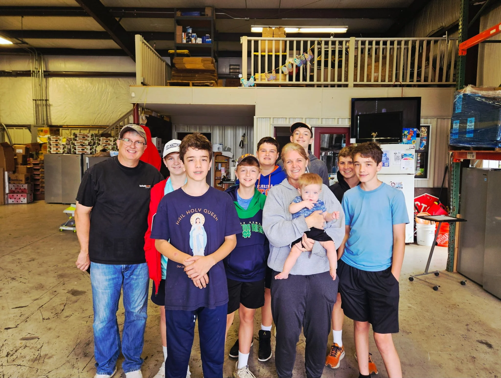 Group of ten people, including seven boys, two women, and one baby, posing together in a warehouse or storage area with shelves, boxes, and equipment in the background.