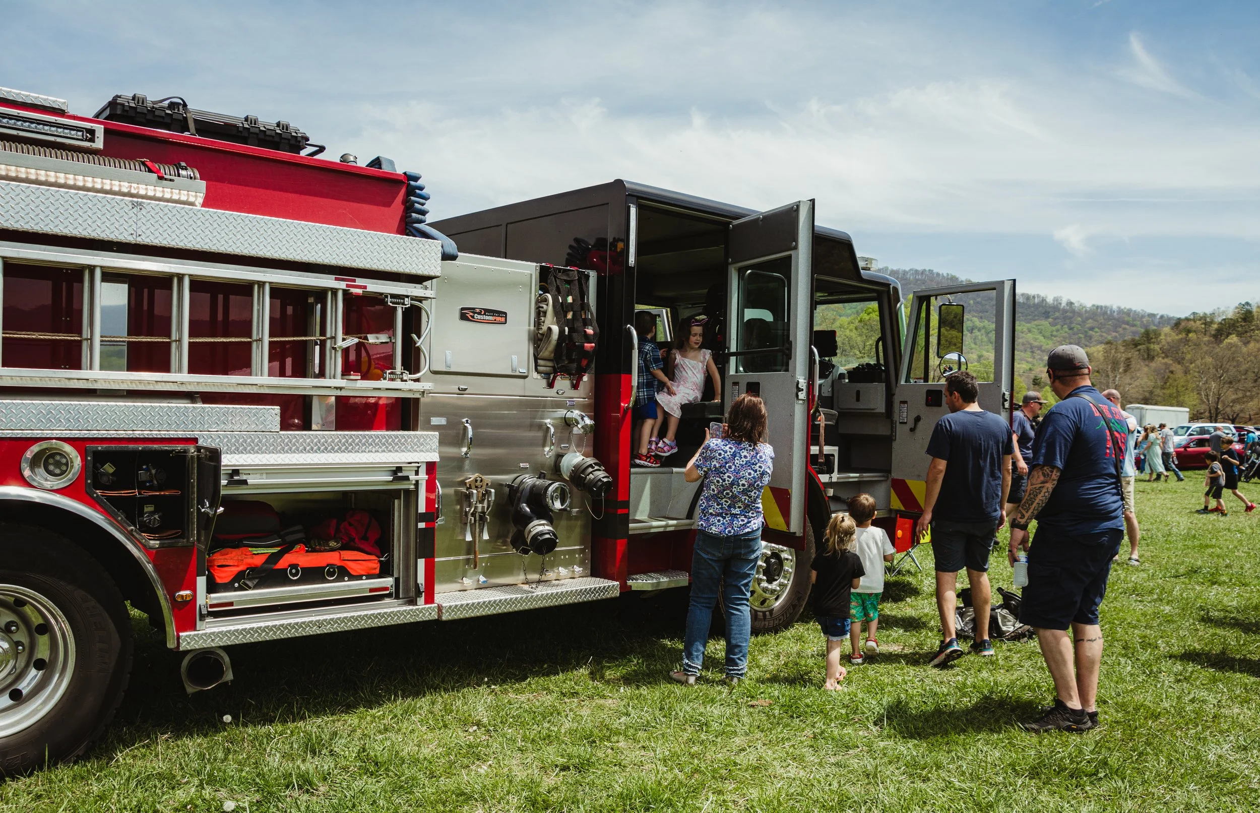 Children and adults gather around a fire truck at an outdoor event on a sunny day, with green hills and a blue sky in the background.