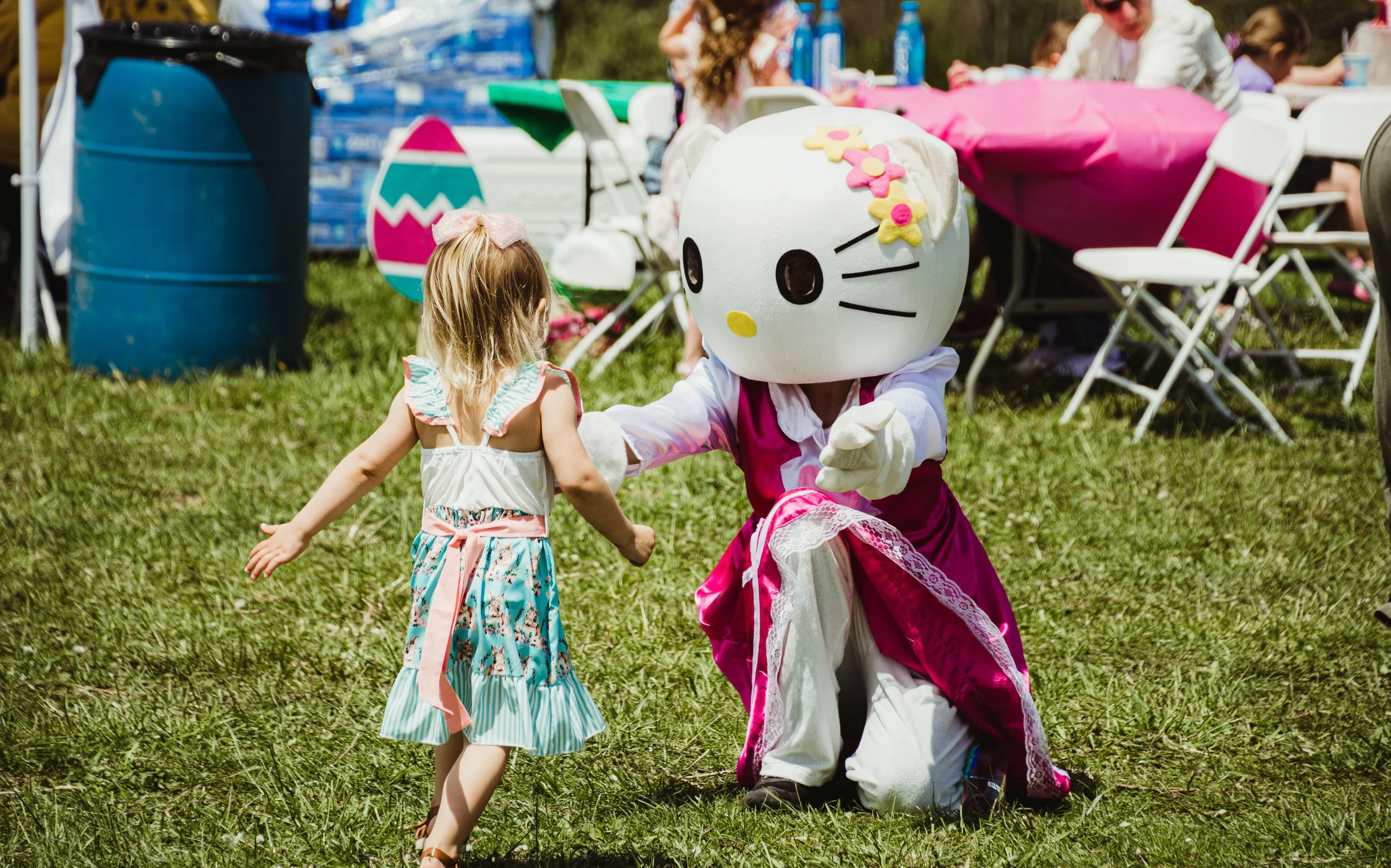 A person in a Hello Kitty costume kneels on the grass, holding hands with a young girl at an outdoor event. The girl is wearing a colorful dress with ruffled sleeves and sandals. In the background, there are tables, chairs, and other people, indicati