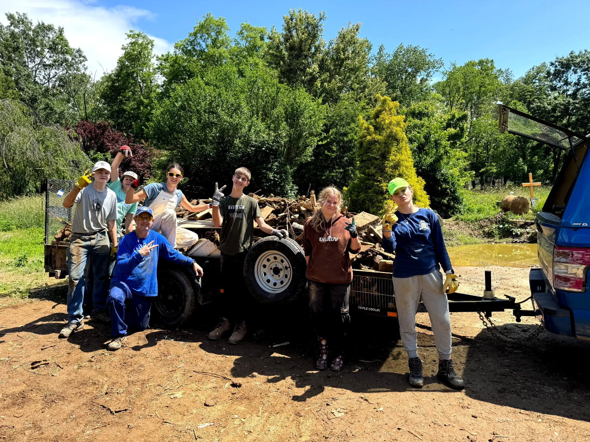 Group of people working outdoors on a sunny day, standing and sitting near a trailer loaded with debris, with trees and a pond in the background.