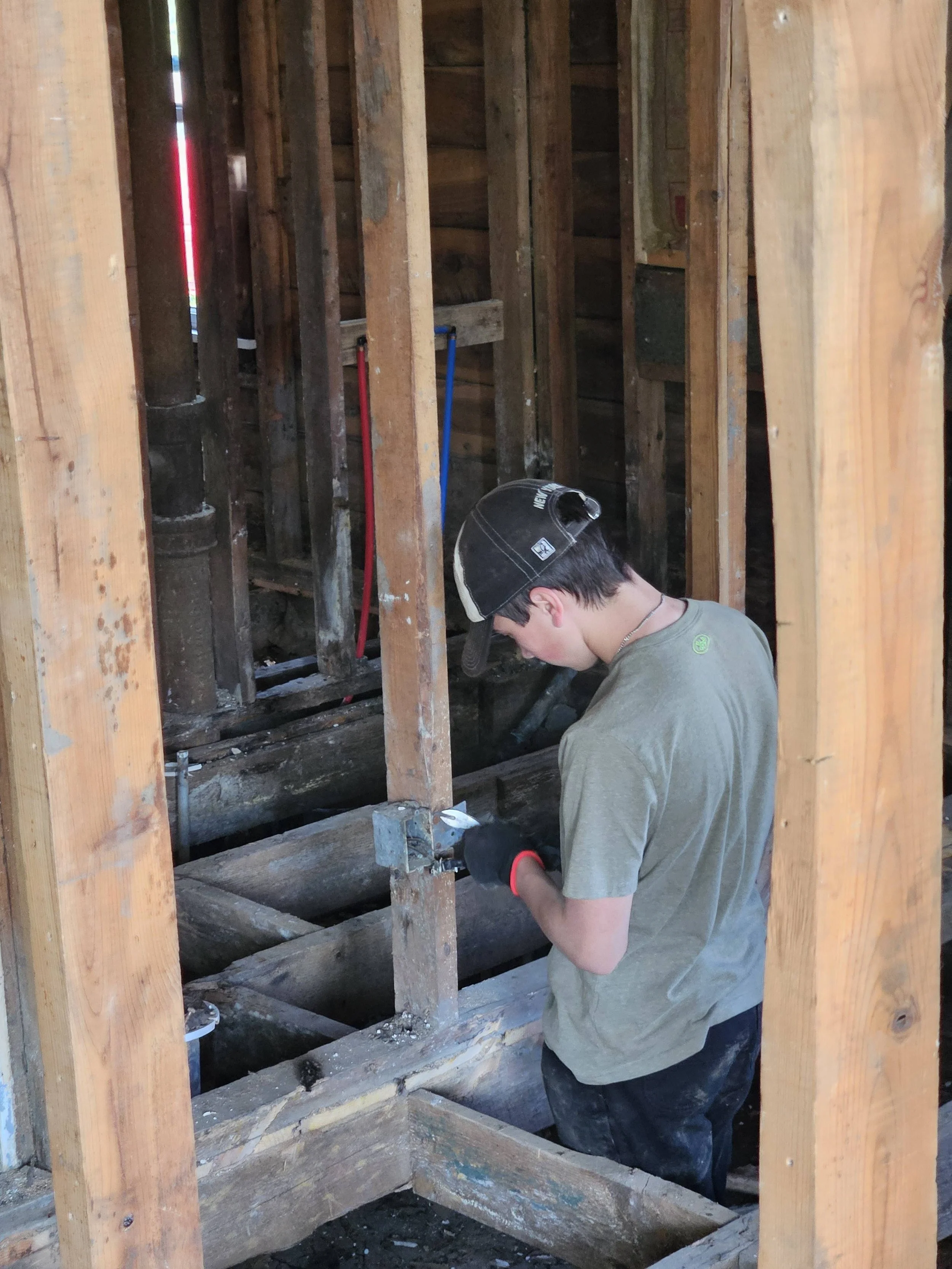 A young person wearing a black cap, gloves, and a beige t-shirt working with tools inside a wooden frame of a house renovation project.