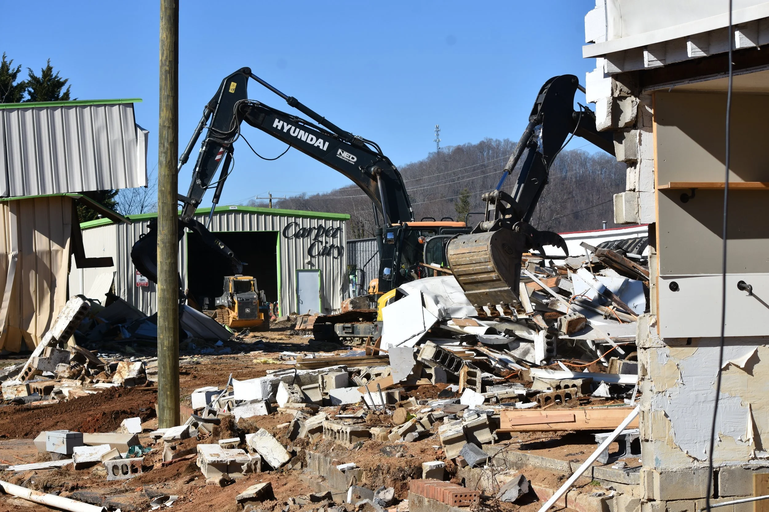 Construction site with an excavator demolishing a brick and wood building, debris scattered on the ground, metal and wood structures, and industrial buildings in the background.