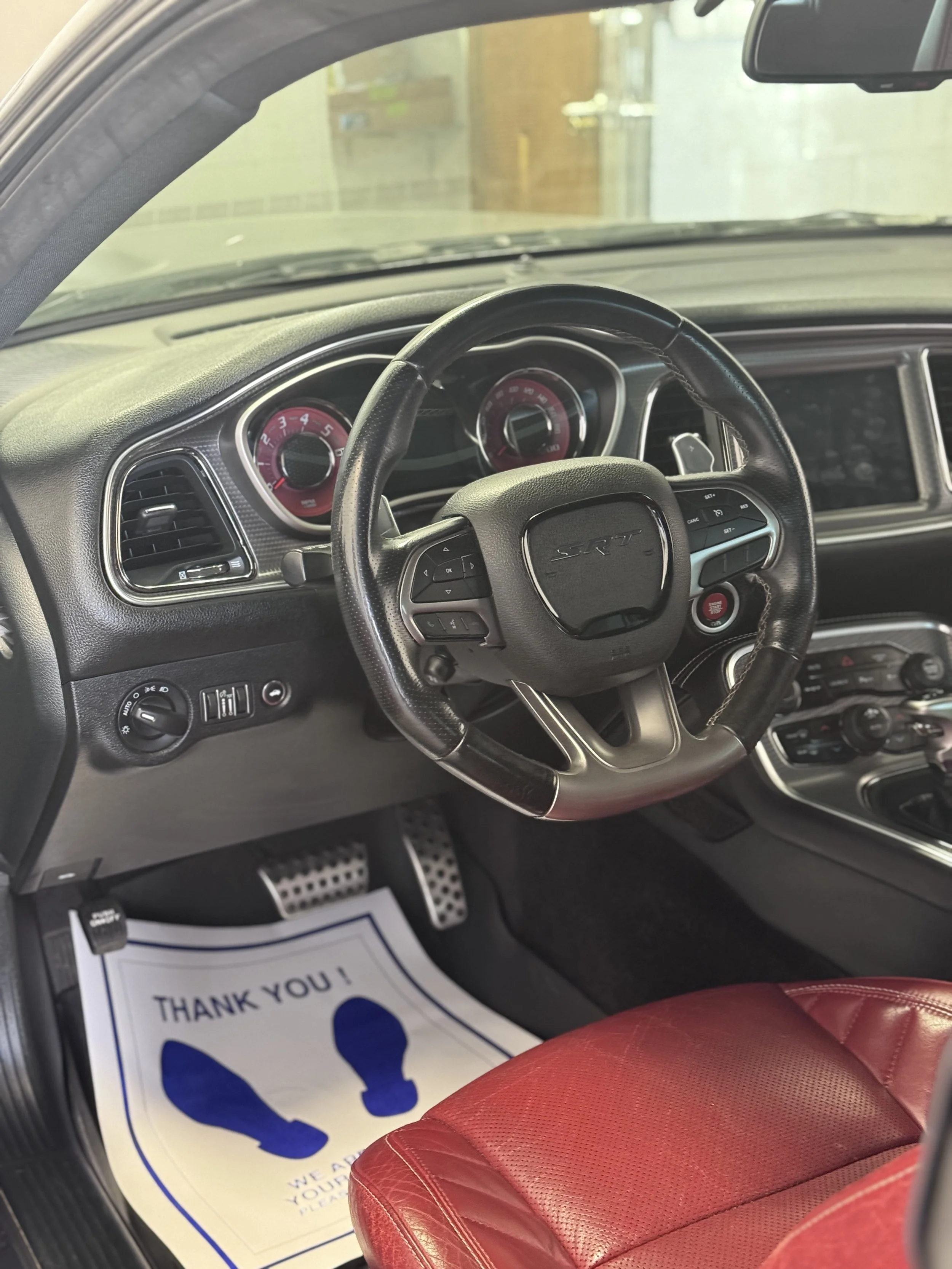 View of a car's interior, showing the steering wheel, dashboard, red leather seat, and a paper floor mat with blue footprints and the phrase 'Thank You' on the floor.