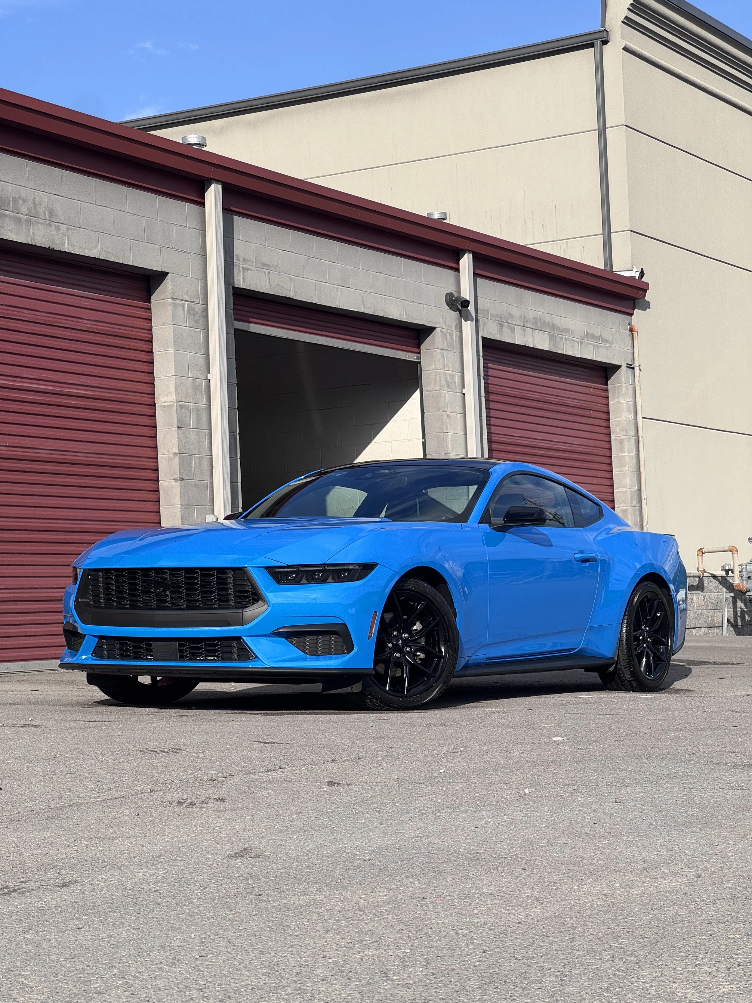 A bright blue sports car parked on a city street in front of a building with red roll-up garage doors and beige walls. The car has black wheels and a sleek design.