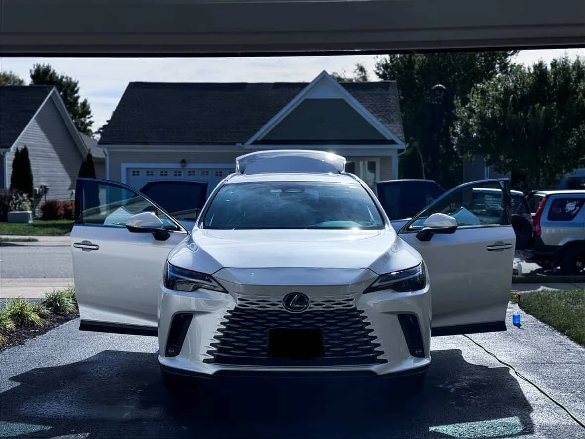 A white Lexus SUV parked in a driveway with its front and side doors open, a house with a driveway and a garage in the background, and a water bottle on the ground to the right.