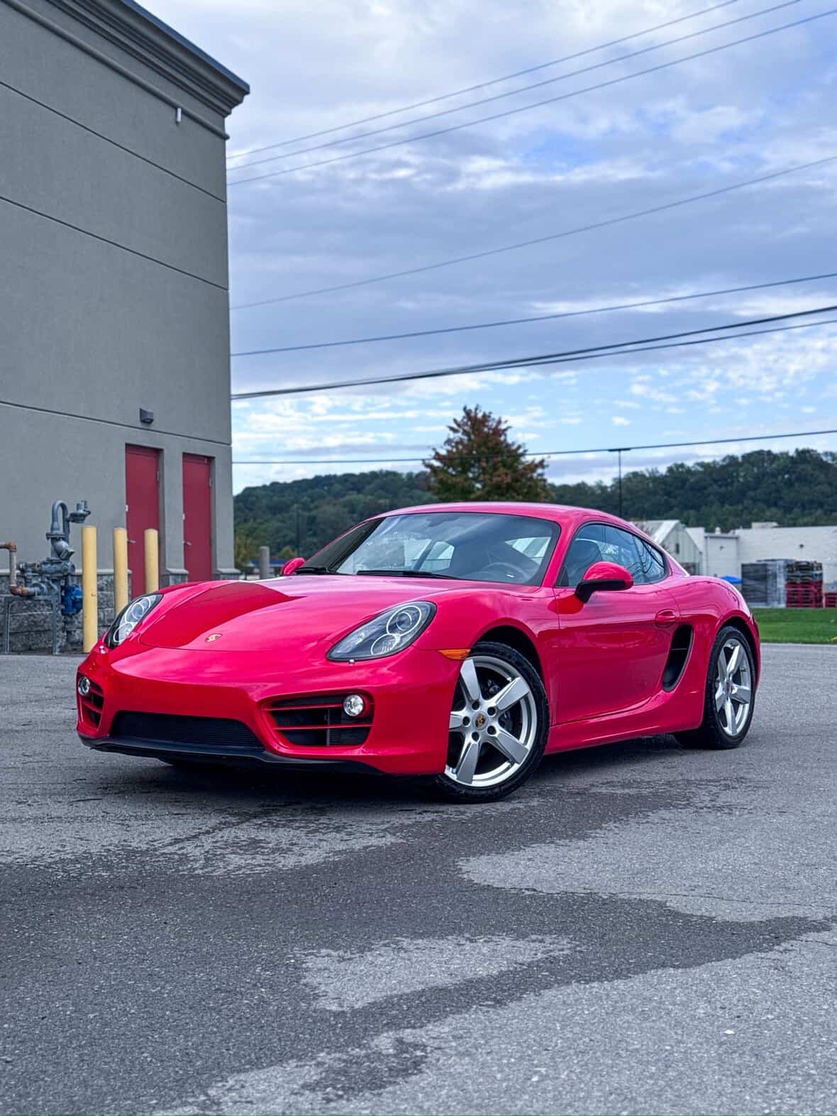 A red Porsche sports car parked outdoors near a building with red doors and a grassy area with trees in the background.