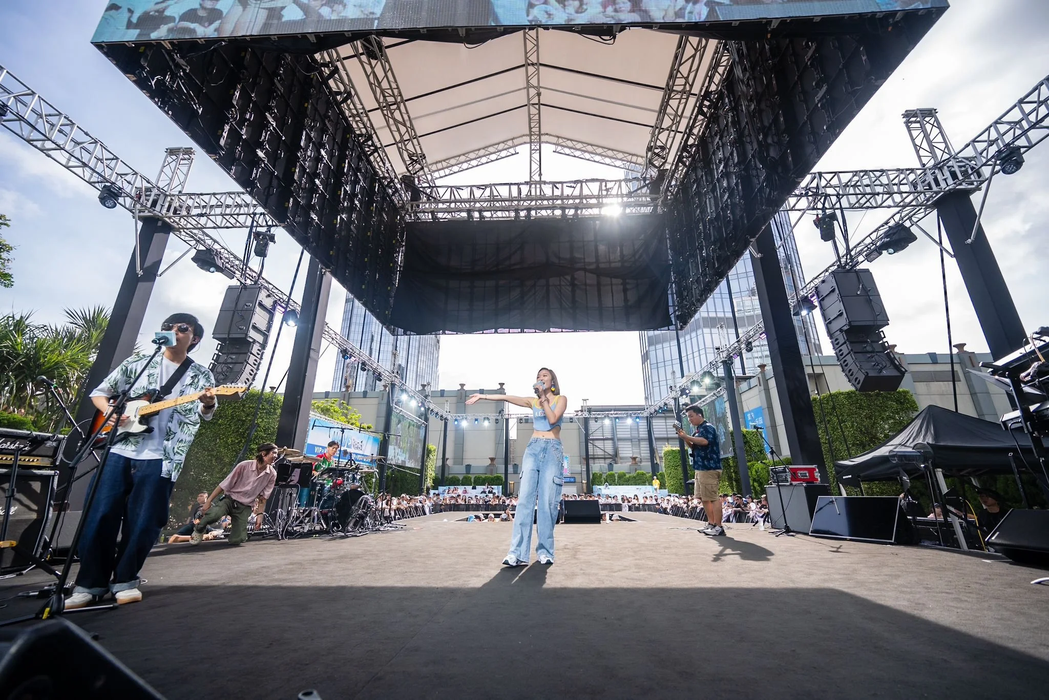 A band performing on an outdoor stage with a singer, guitarist, and drummer. Lights and speakers are set up above the stage, with an audience in front.