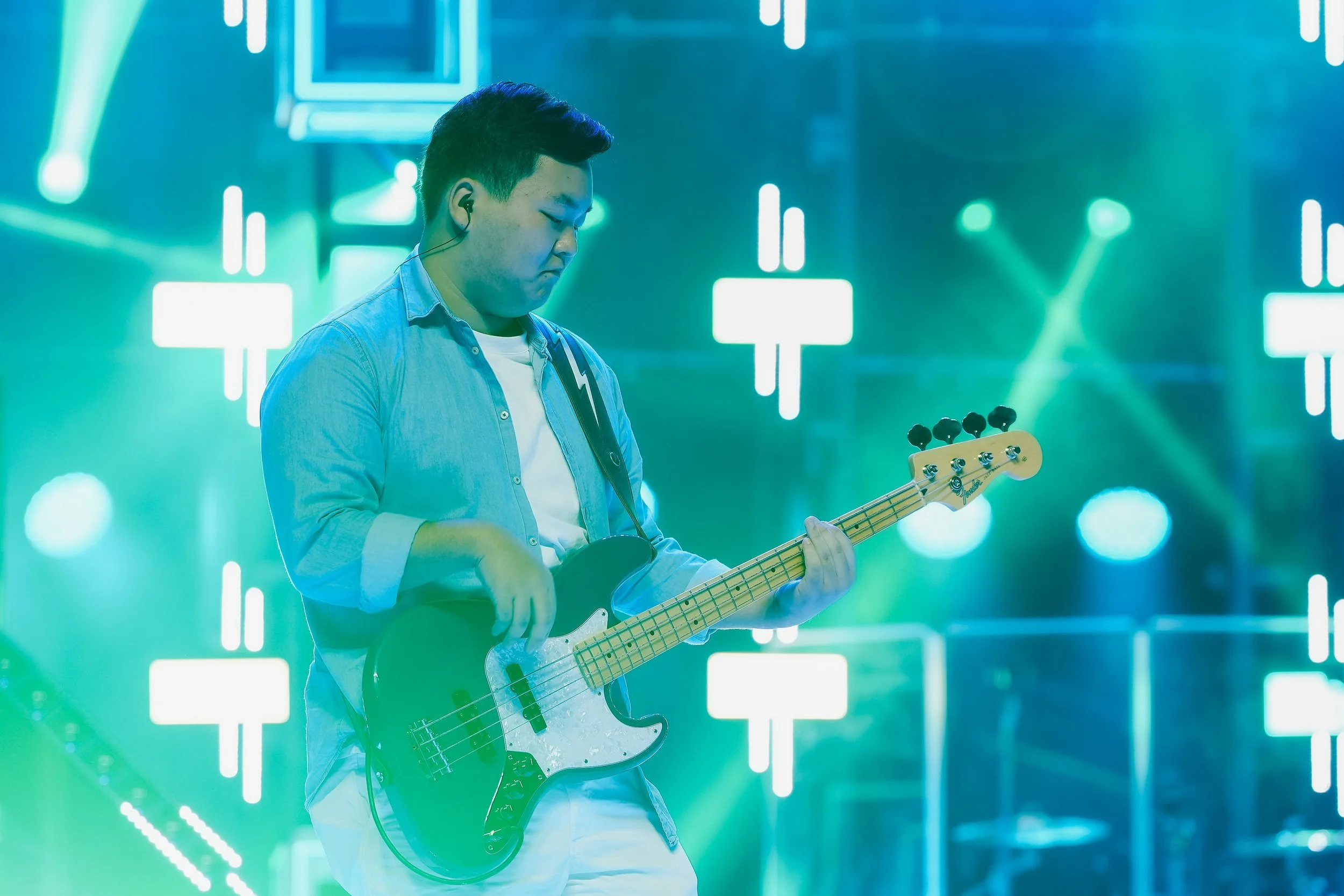 A young man playing an electric bass guitar on stage with a vibrant light display in the background.