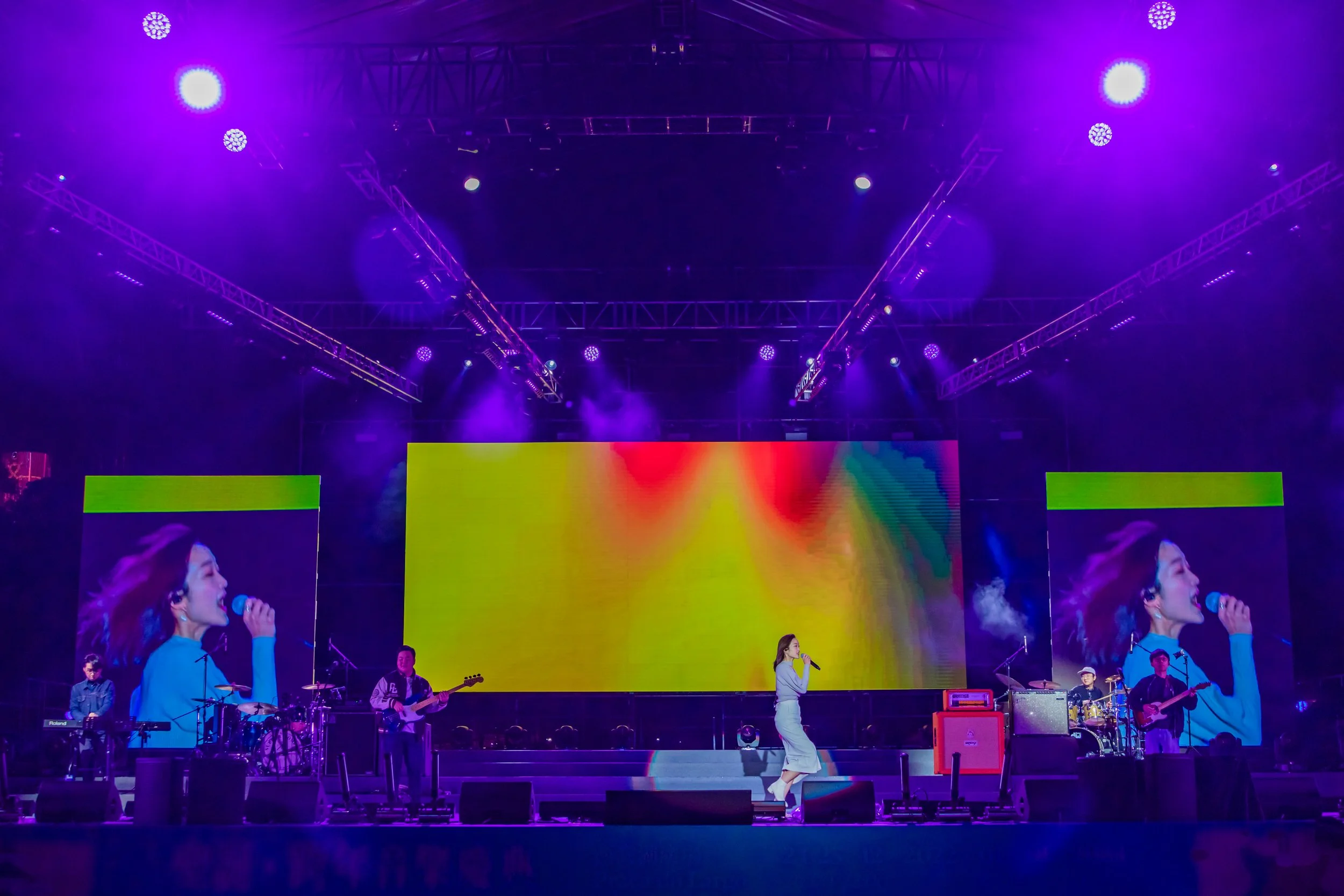 A live concert stage with a female singer in white pants and top holding a microphone, surrounded by a band with keyboards, guitars, and drums, under purple stage lights, with a large colorful LED screen in the background