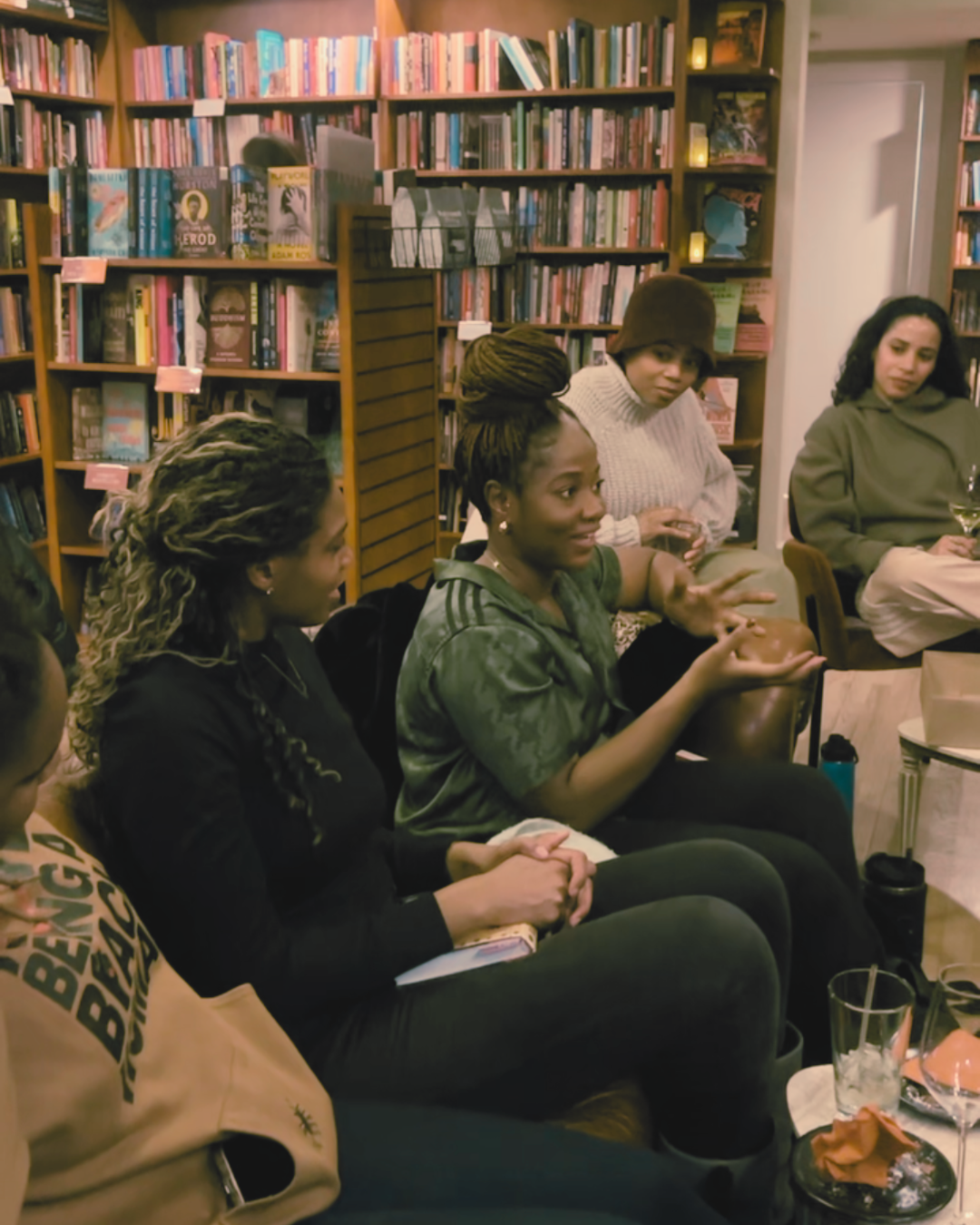 A group of women sitting and talking in a cozy, well-stocked bookstore or library, with bookshelves filled with colorful books behind them.
