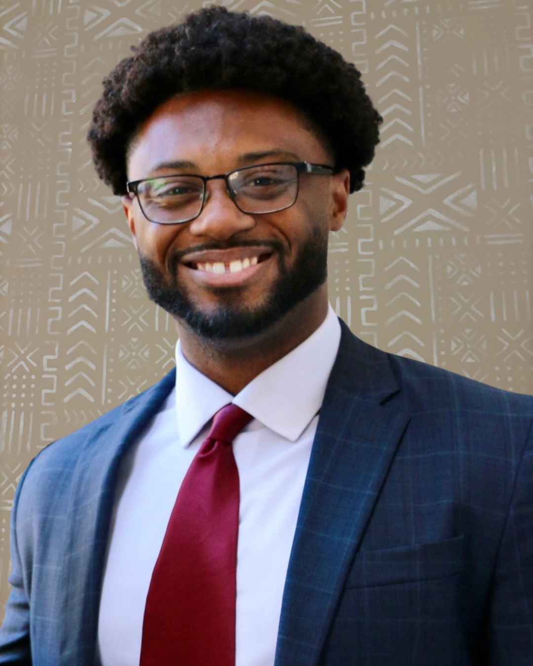 A smiling man with glasses and a beard, dressed in a checked blue suit, white shirt, and maroon tie, standing outdoors in front of a neutral background.