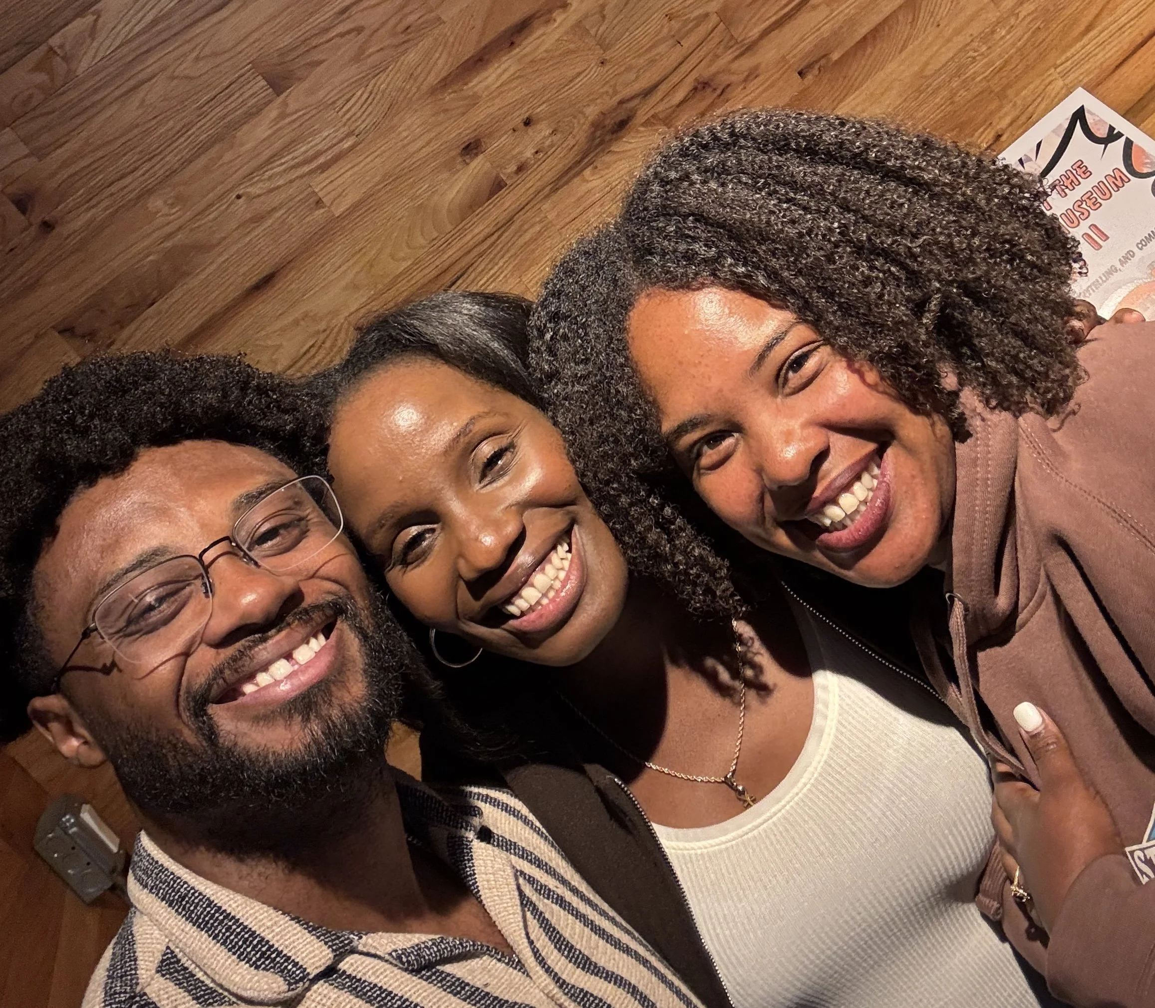 Close-up photo of three people women, side by side, with wooden wall background. The man wears glasses, one woman has straight hair, and the third has curly hair They are wearing casual clothing.